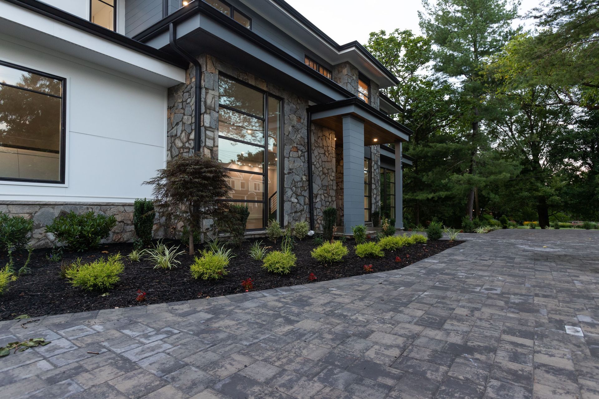 Modern house with stone facade, large windows, landscaped driveway, and lush greenery.