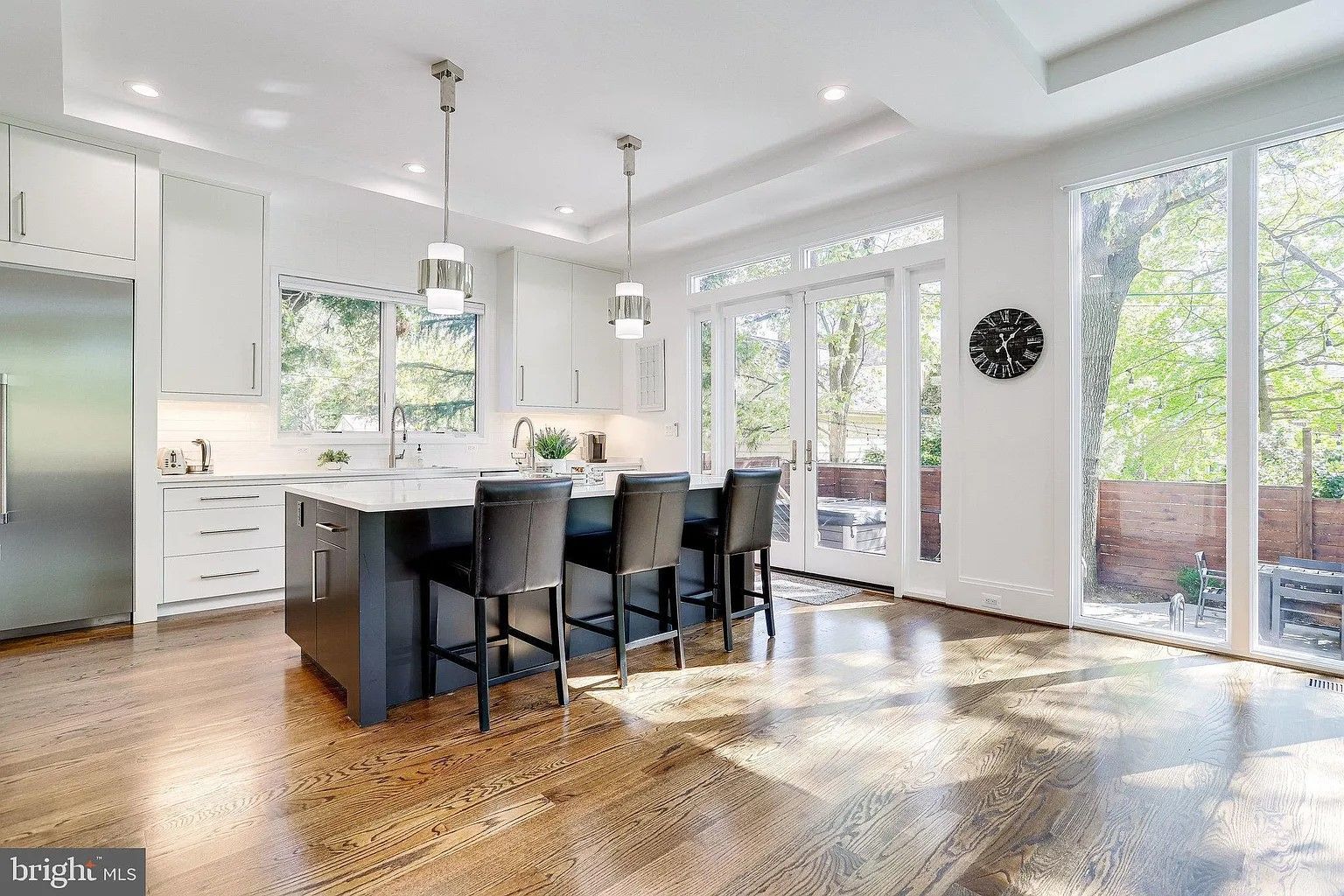 Modern white kitchen with dark island and stools, large windows looking out at trees.