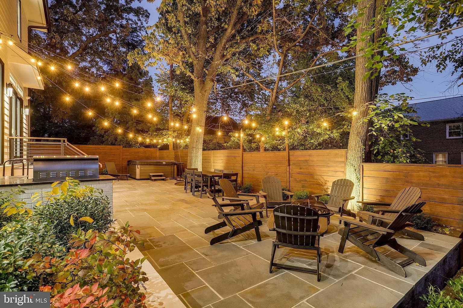 Patio with string lights, chairs around a fire pit, trees, and greenery. Dusk setting.