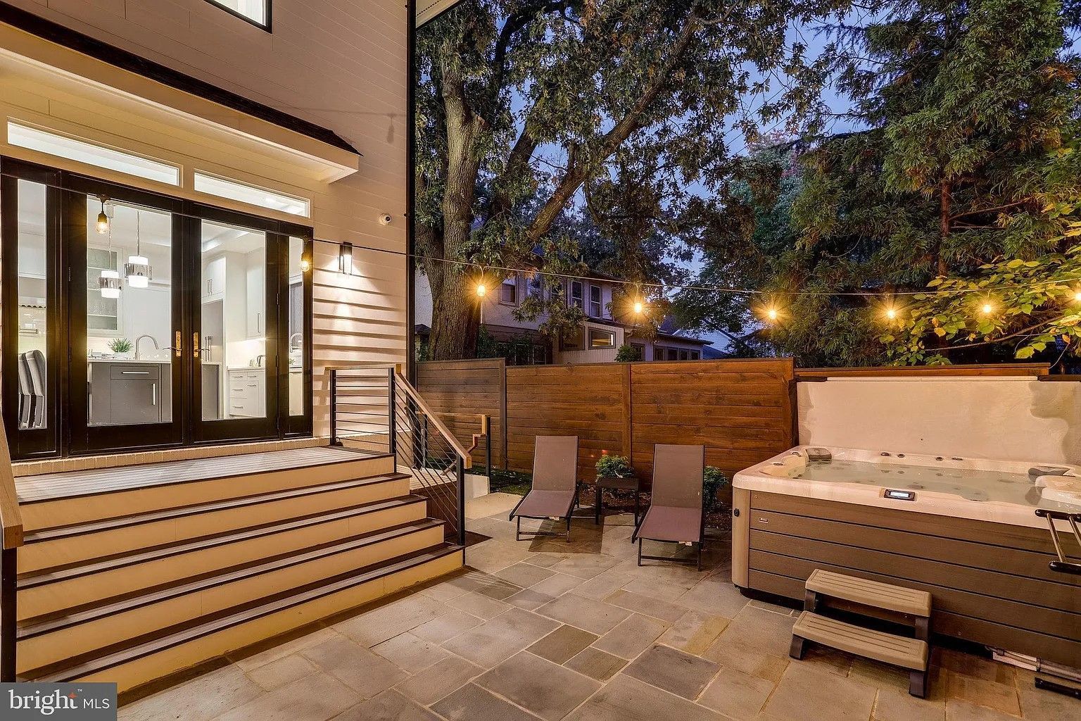 Patio with hot tub, chairs, and string lights next to house with glass doors.