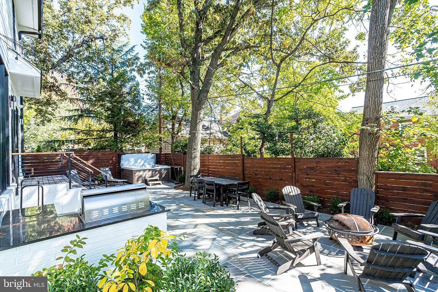 Backyard with a grill, seating, a hot tub, and a wooden fence surrounded by trees.