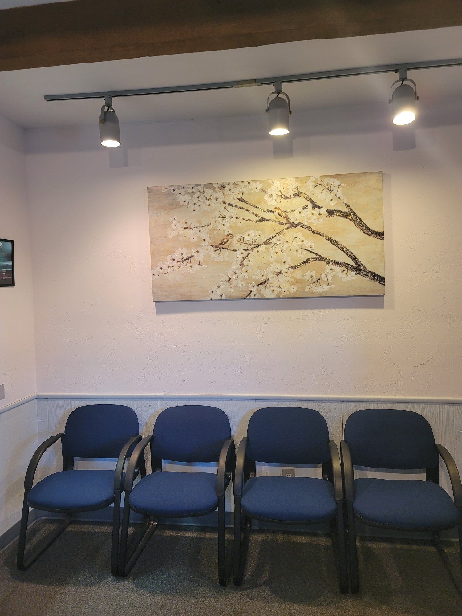 A row of blue chairs in a waiting room with a painting on the wall above them