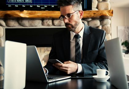 A professional in a suit and tie sits at a desk with a laptop and phone, in front of a stone fireplace with a screen.