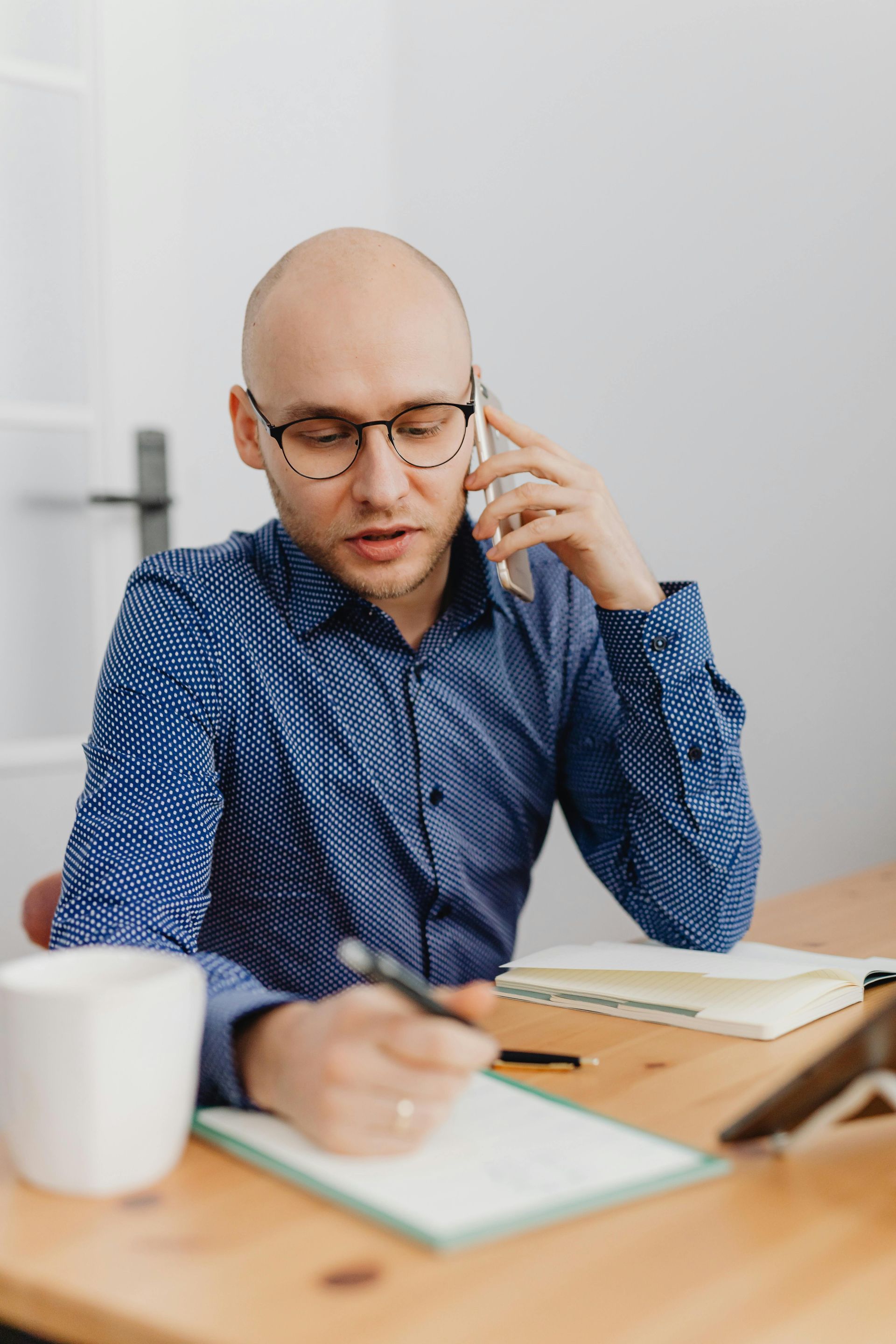 A person in a blue patterned shirt talks on a phone while writing in a notebook at a desk.