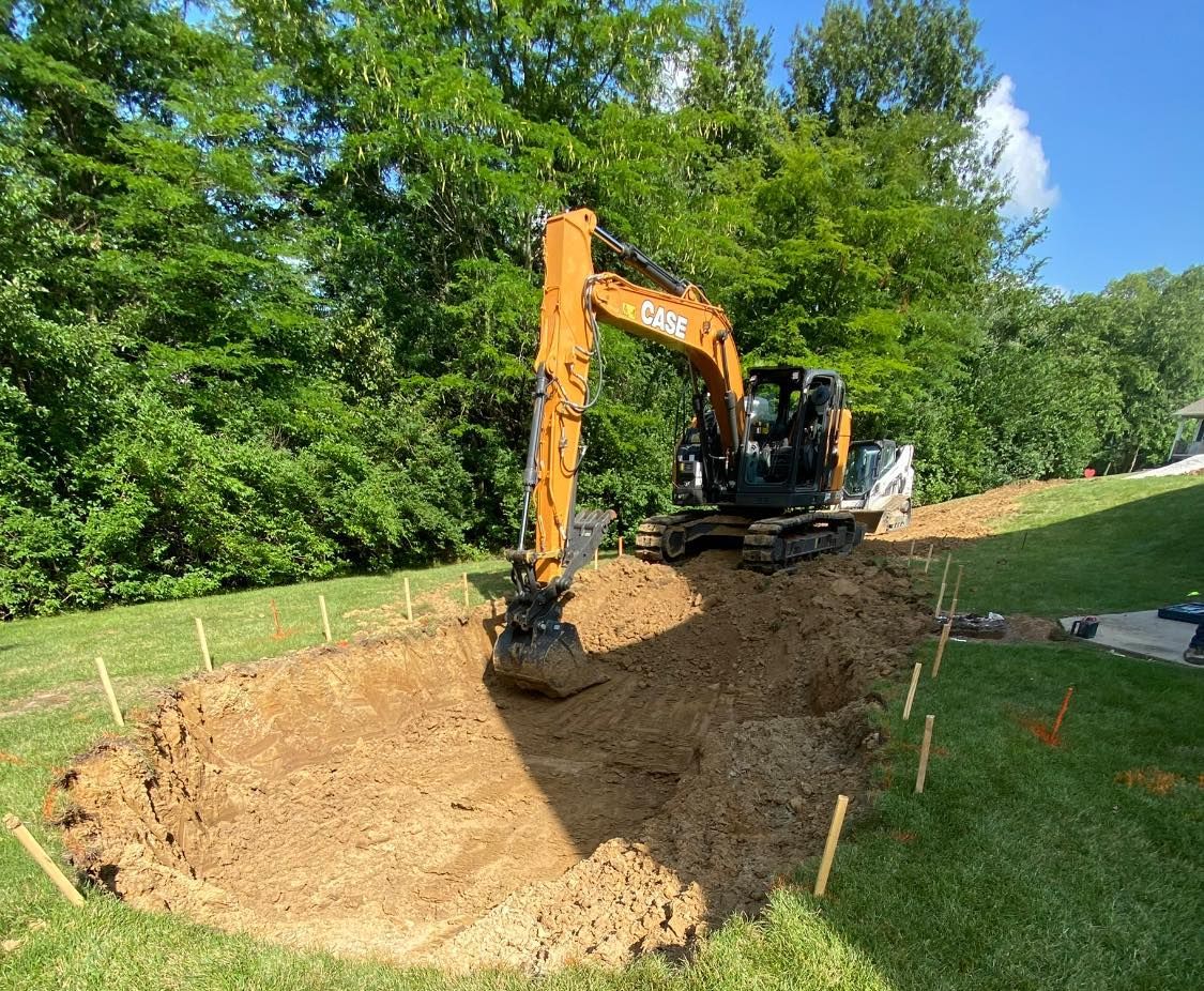 An excavator digs a rectangular hole in a grassy backyard, surrounded by wooden stakes, trees, and blue sky.