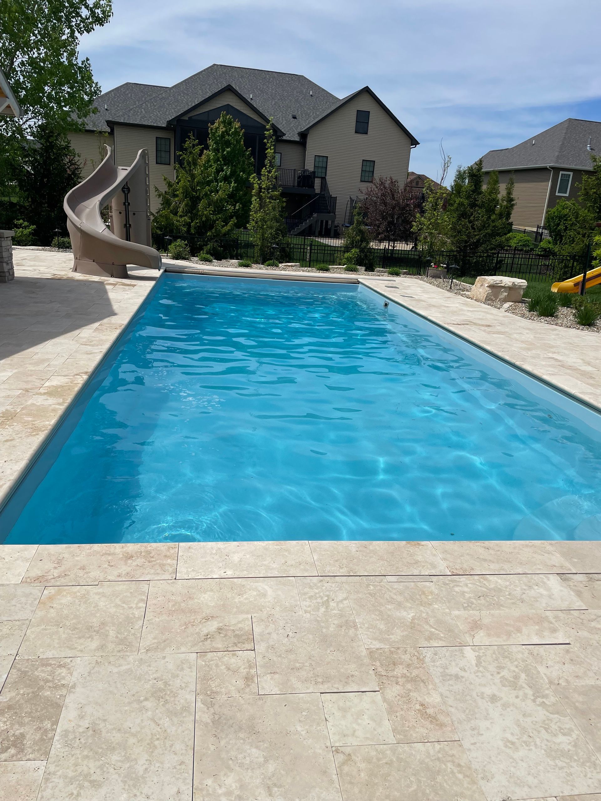 Rectangular pool with turquoise water, beige stone surround, and a house with a slide in the background.