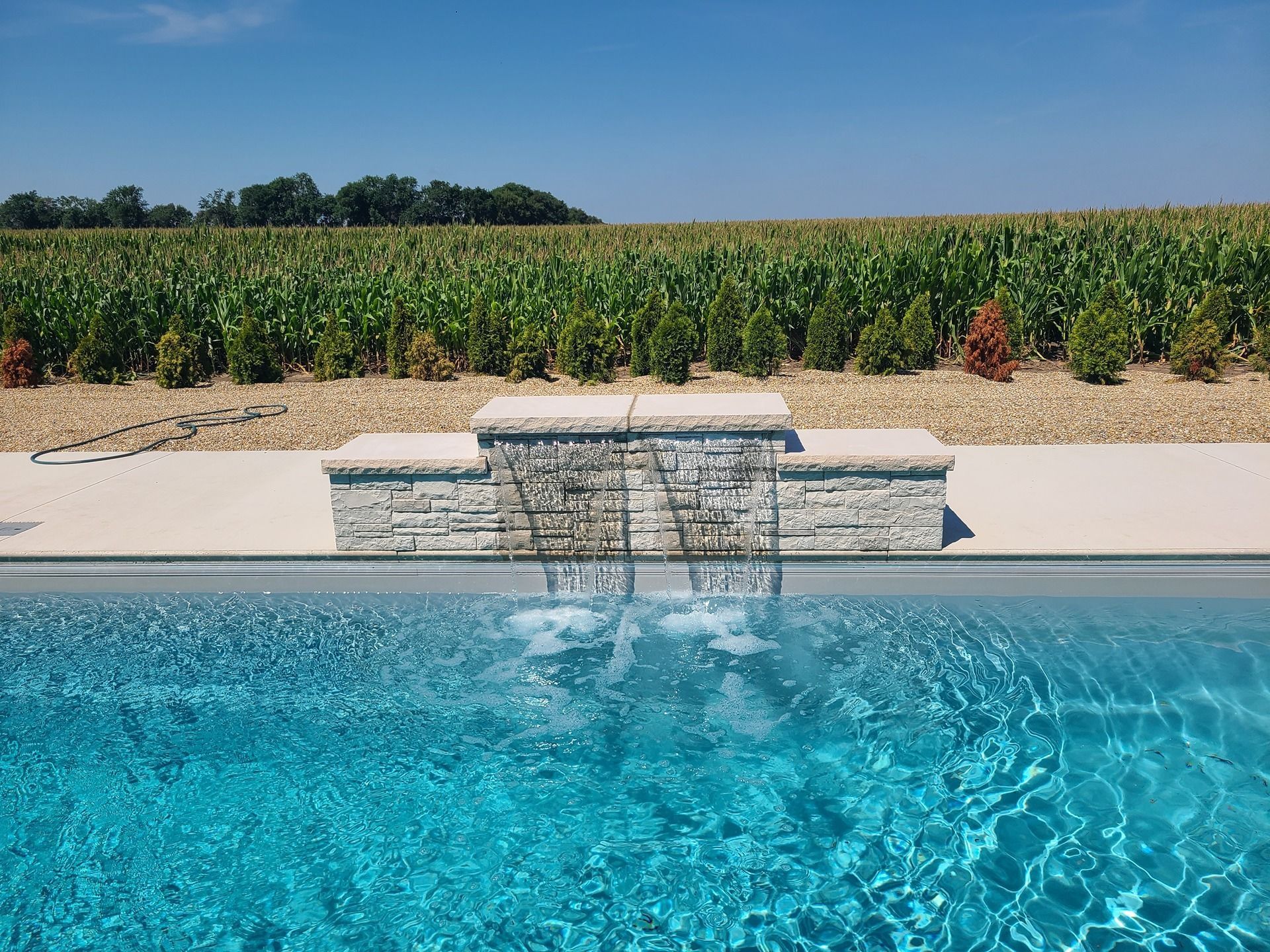 Swimming pool with a stone waterfall feature, reflecting a blue sky and a field of crops.