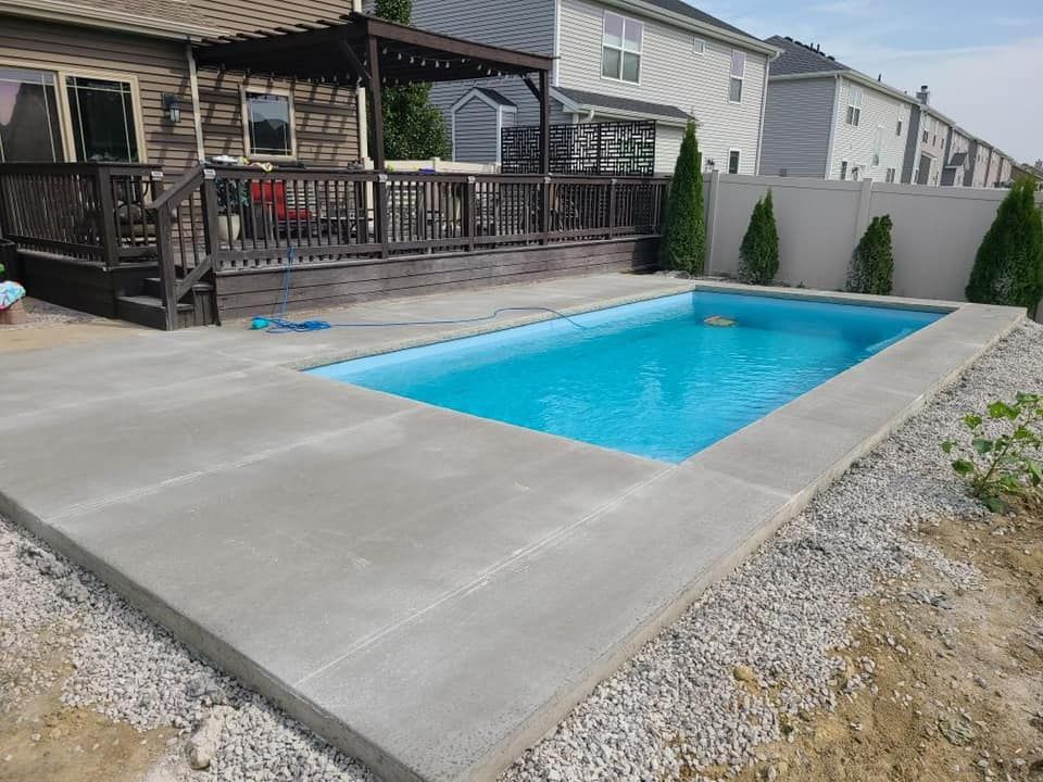 Concrete pool deck surrounding a rectangular pool with turquoise water. A wooden deck and house are in the background.