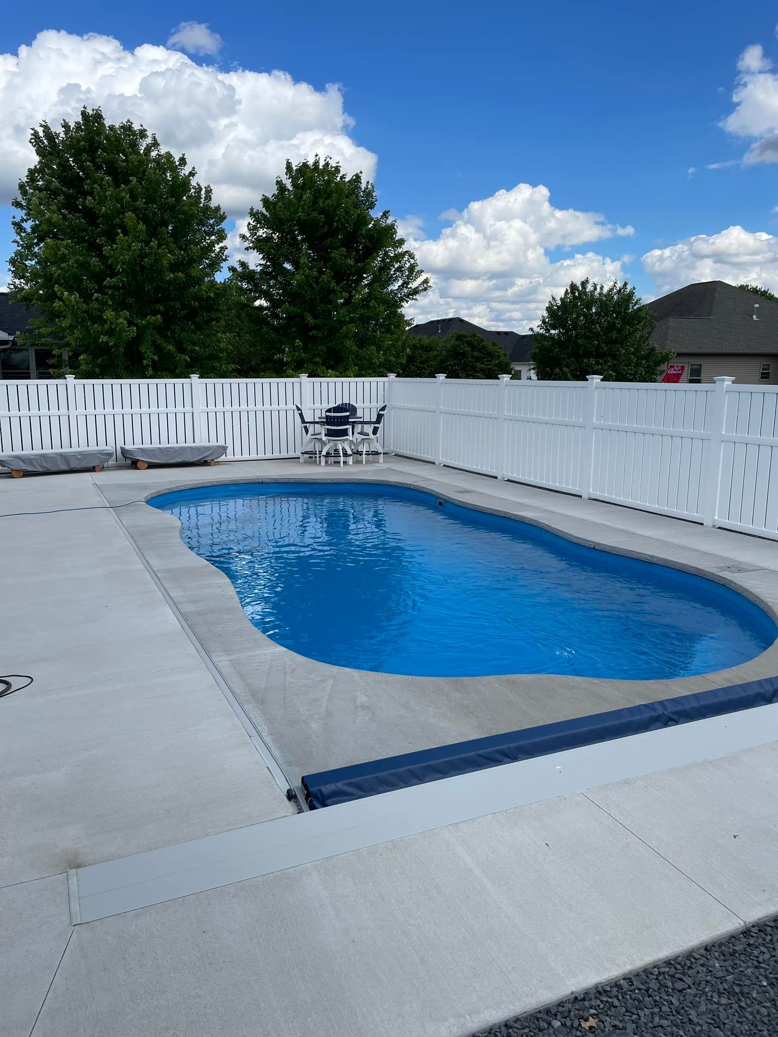 Oval-shaped in-ground pool surrounded by concrete and white fence under a partly cloudy sky.