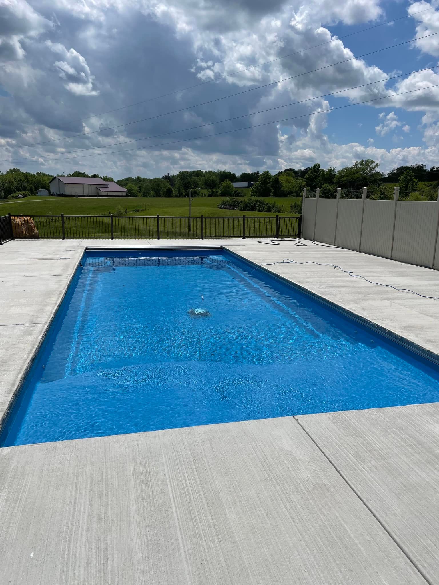 Rectangular blue pool surrounded by concrete, in a grassy field under a cloudy sky.