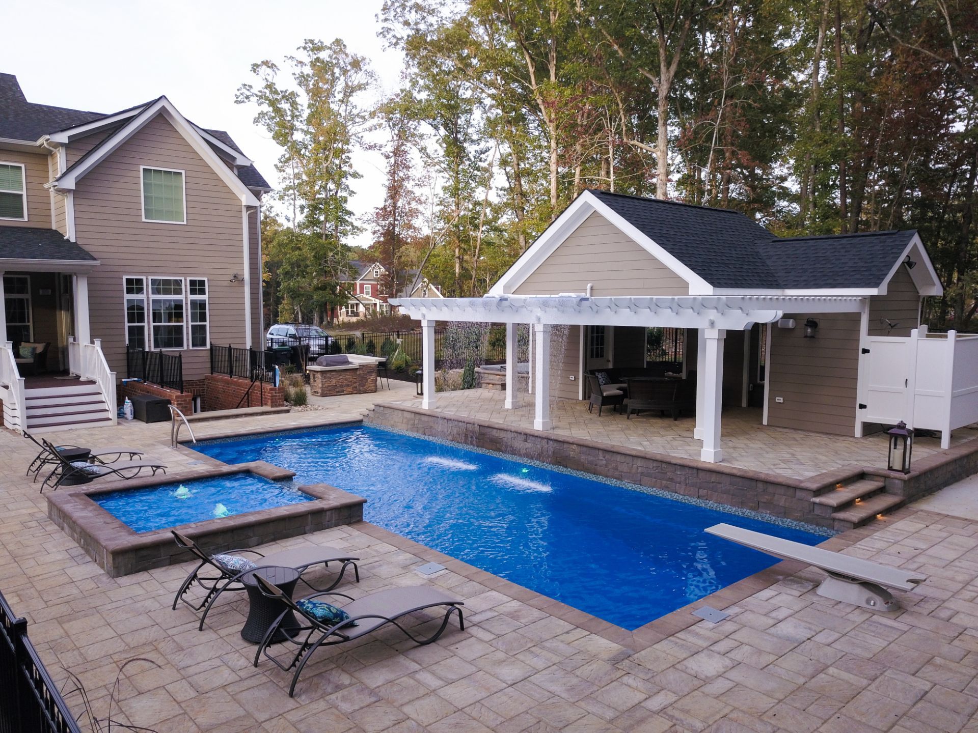 Backyard pool with spa, lounge chairs, patio, and pergola near a house and trees.