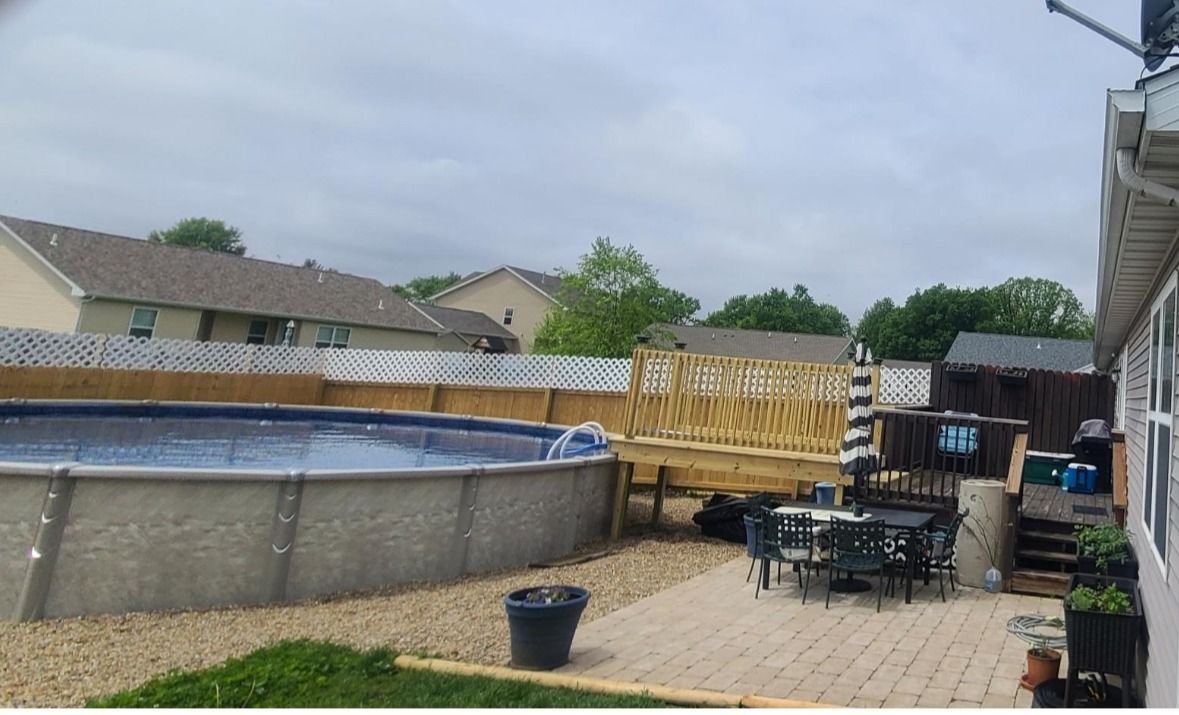 Above-ground pool in a backyard with a wooden deck, patio furniture, and a fence. Overcast sky.