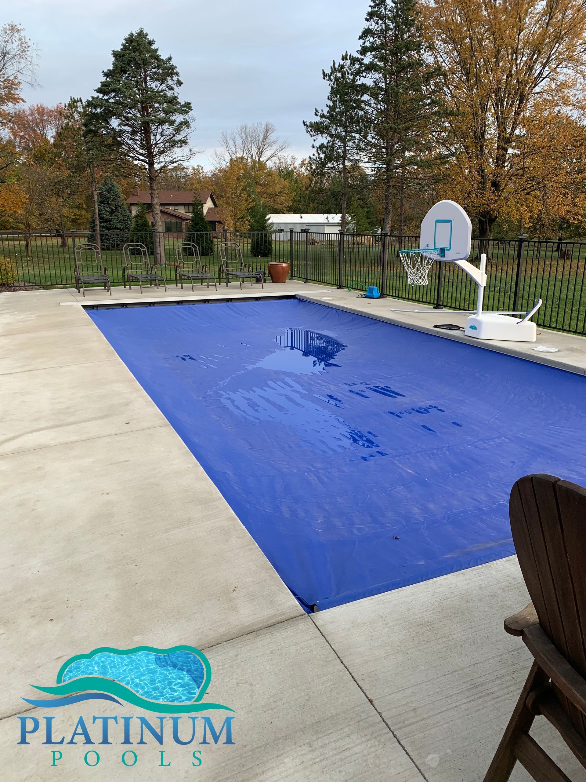 Blue pool cover on a rectangular pool surrounded by concrete. Basketball hoop and trees in the background.