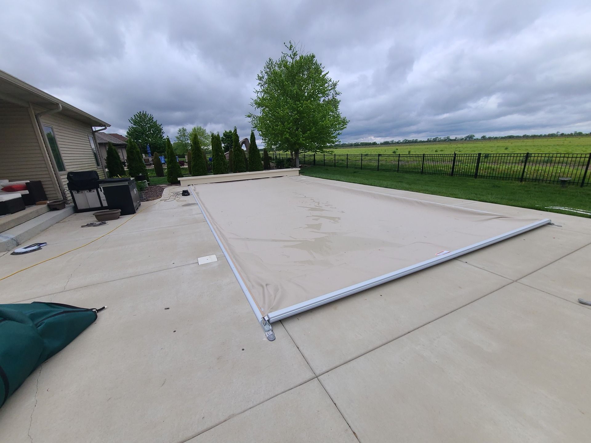 Beige pool cover on a concrete patio, with green foliage and a cloudy sky in the background.