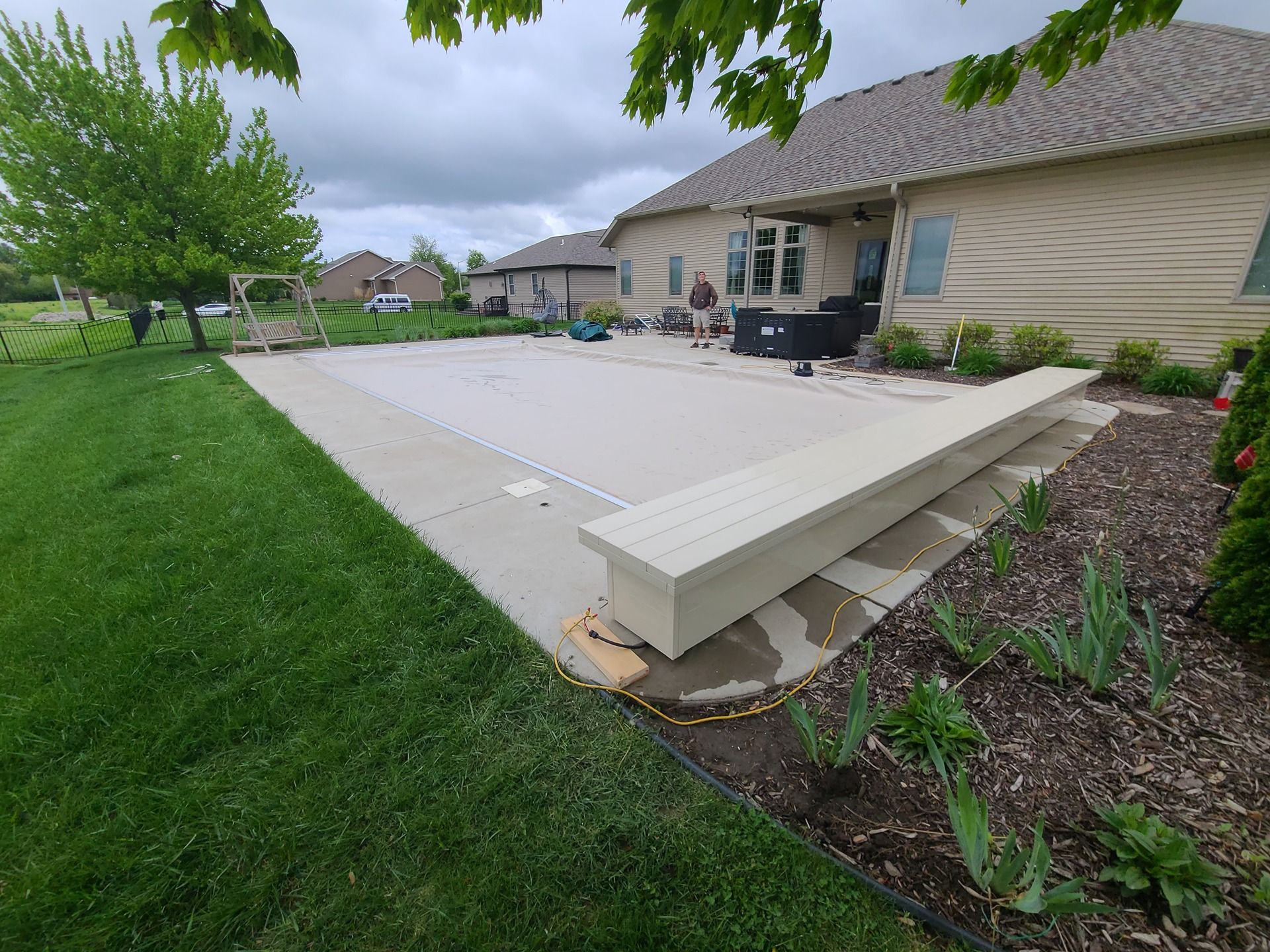 Backyard with a pool cover, concrete patio, and a long bench. Green grass and a cloudy sky.