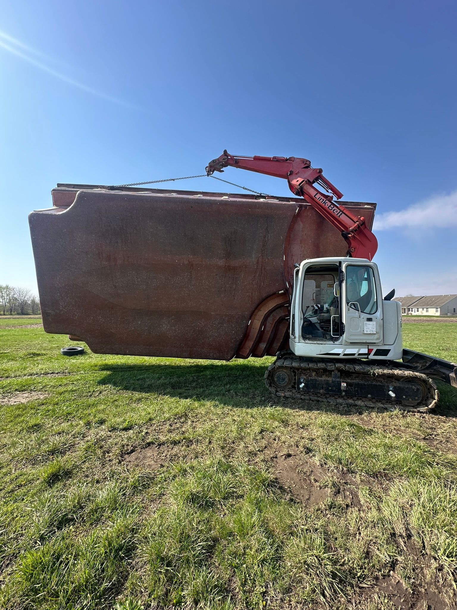 An excavator lifting a large, weathered, rectangular metal structure in a grassy field under a blue sky.