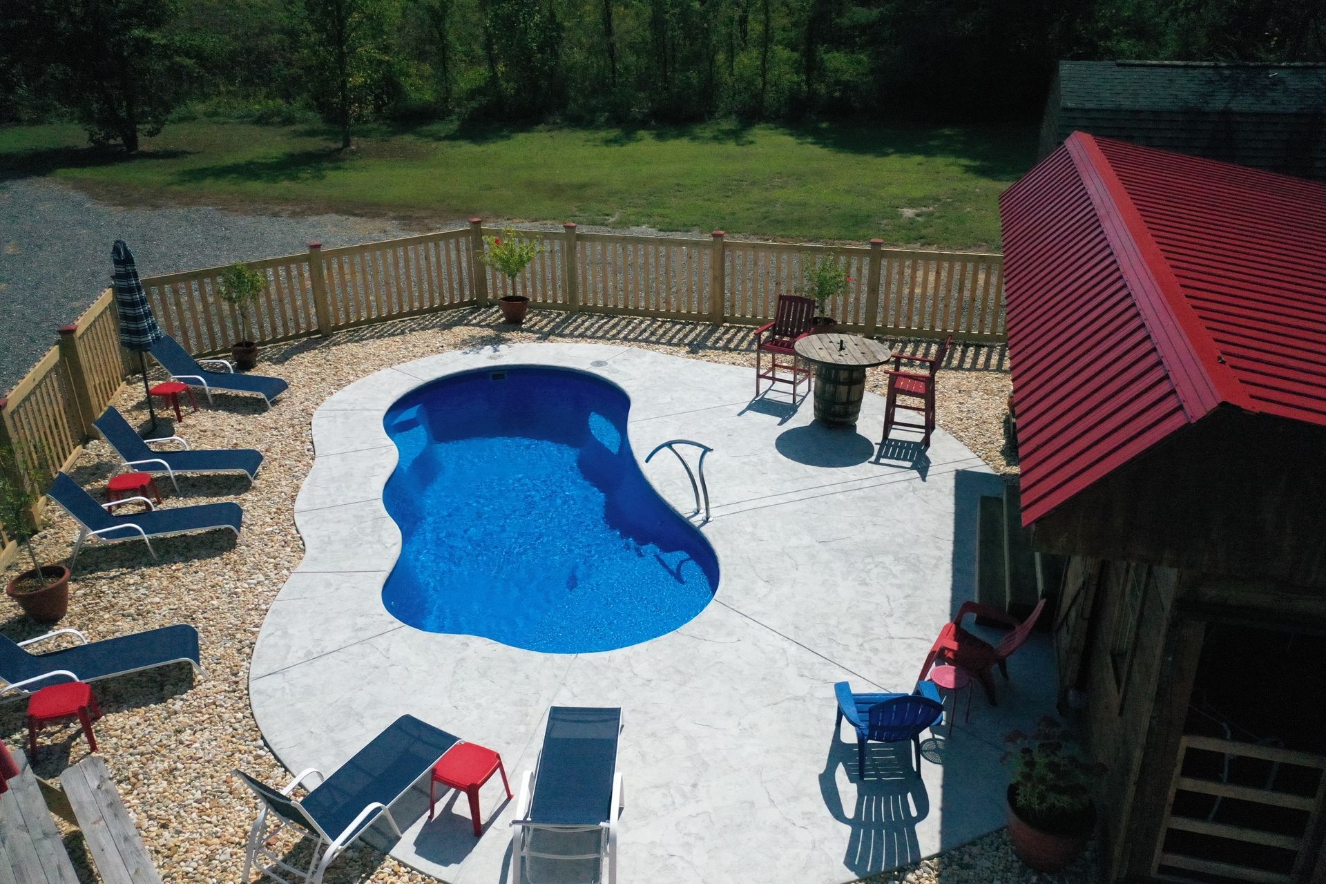 Swimming pool with lounge chairs, fenced patio, and red-roofed building.