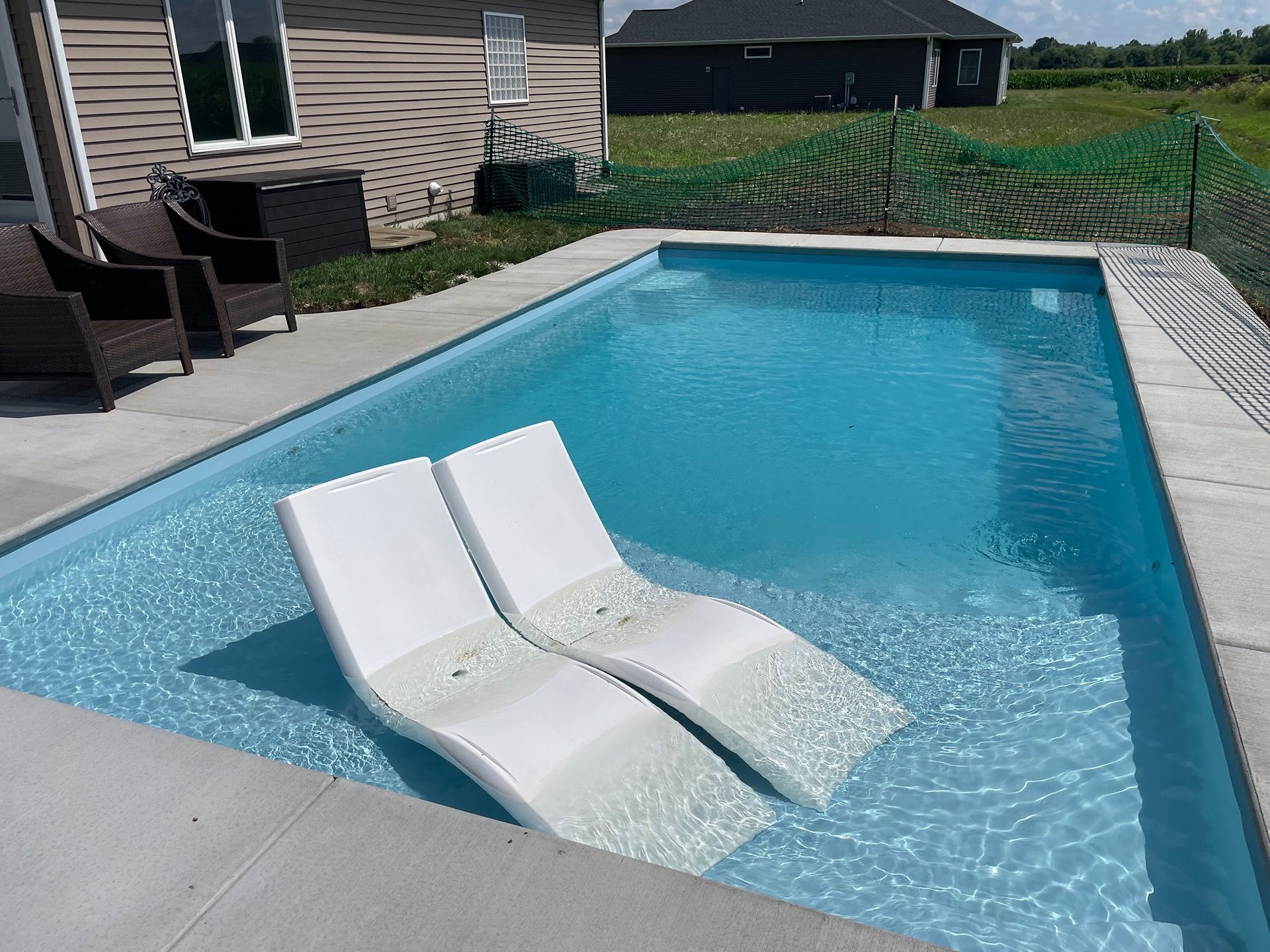 Two white chaise lounge chairs in a rectangular swimming pool with blue water. A house is in the background.