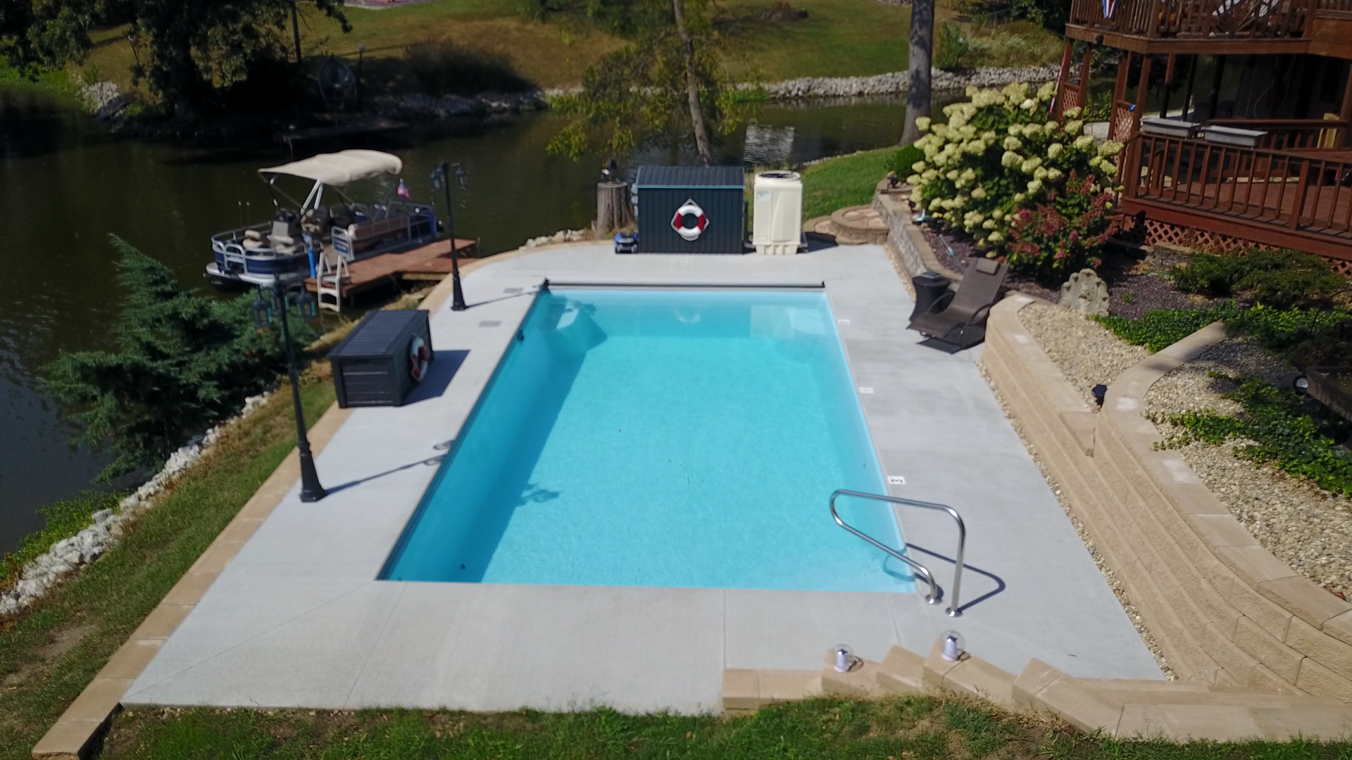 Rectangular pool next to a lake, with a dock and a house. Concrete patio.