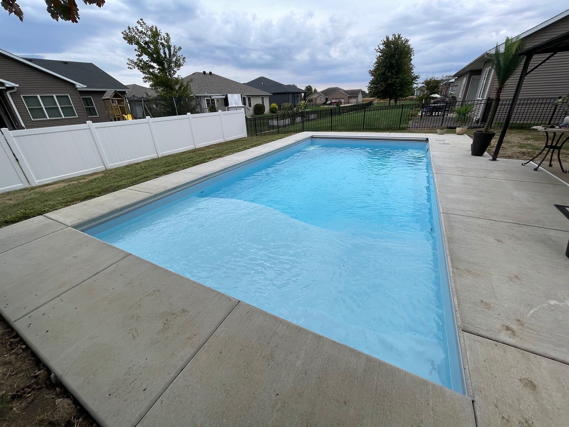 Rectangular swimming pool with light blue water surrounded by concrete, in a backyard setting.