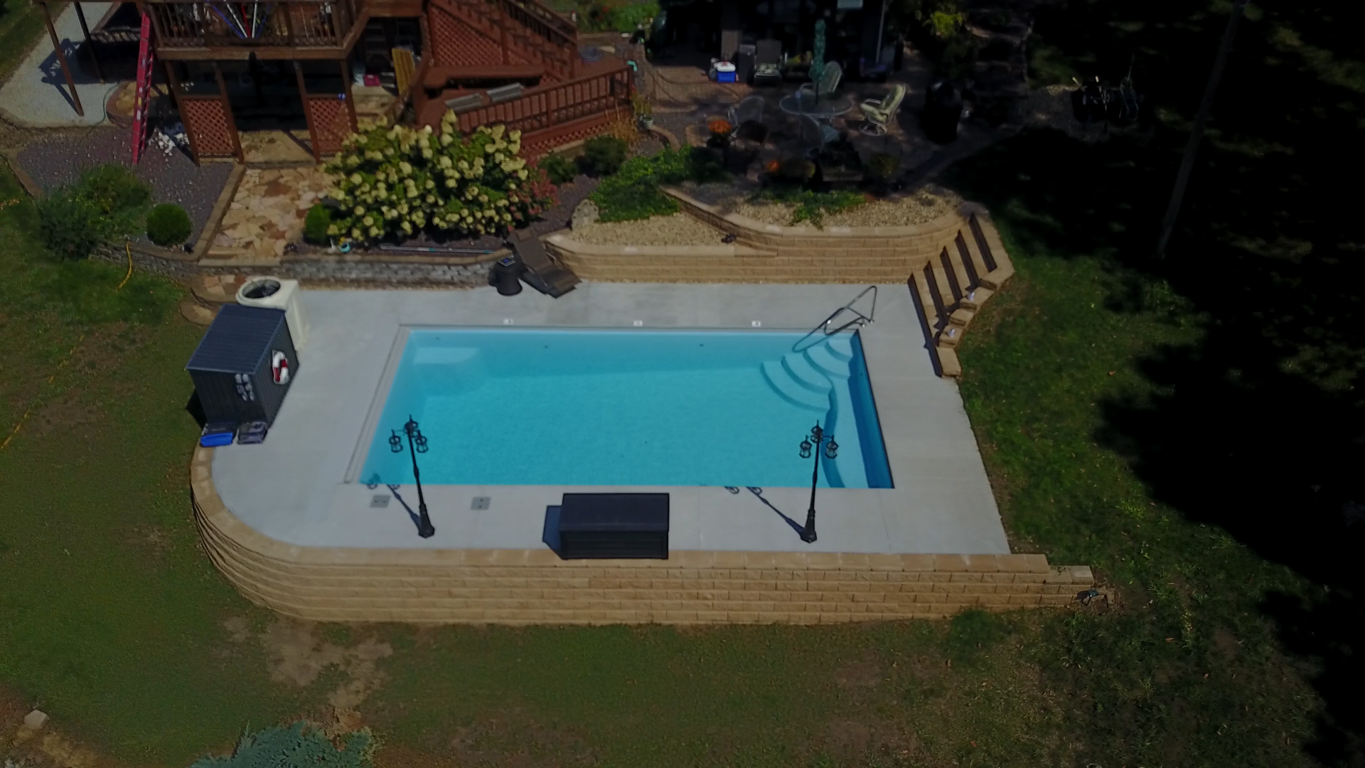 Aerial view of a rectangular pool with a stone retaining wall and steps, next to a multi-level house.