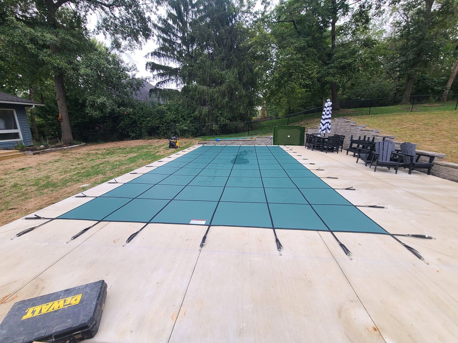 Green pool cover on a concrete patio, surrounded by grass and trees.