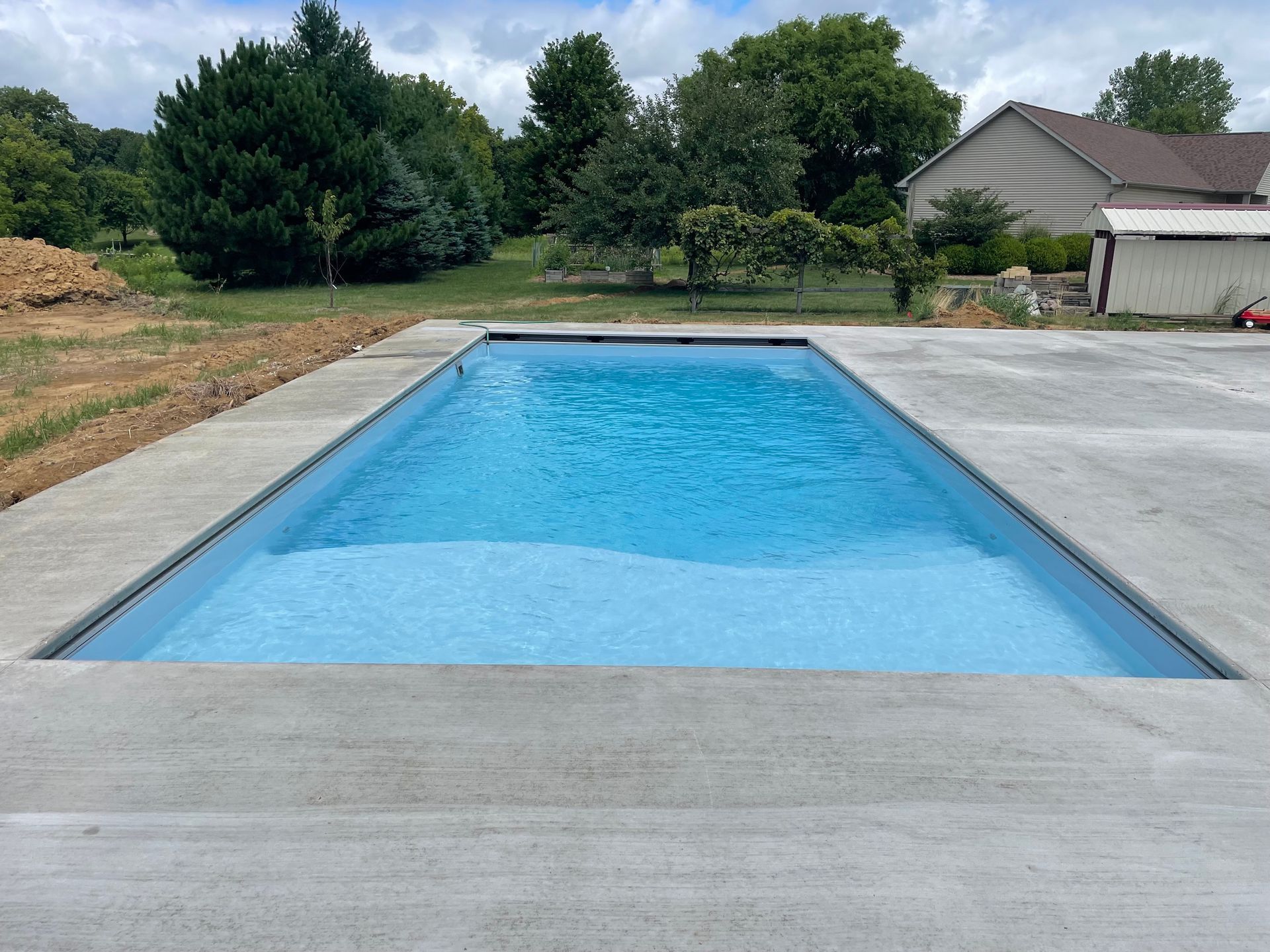 Rectangular swimming pool filled with blue water surrounded by concrete, in a yard with trees and a house.