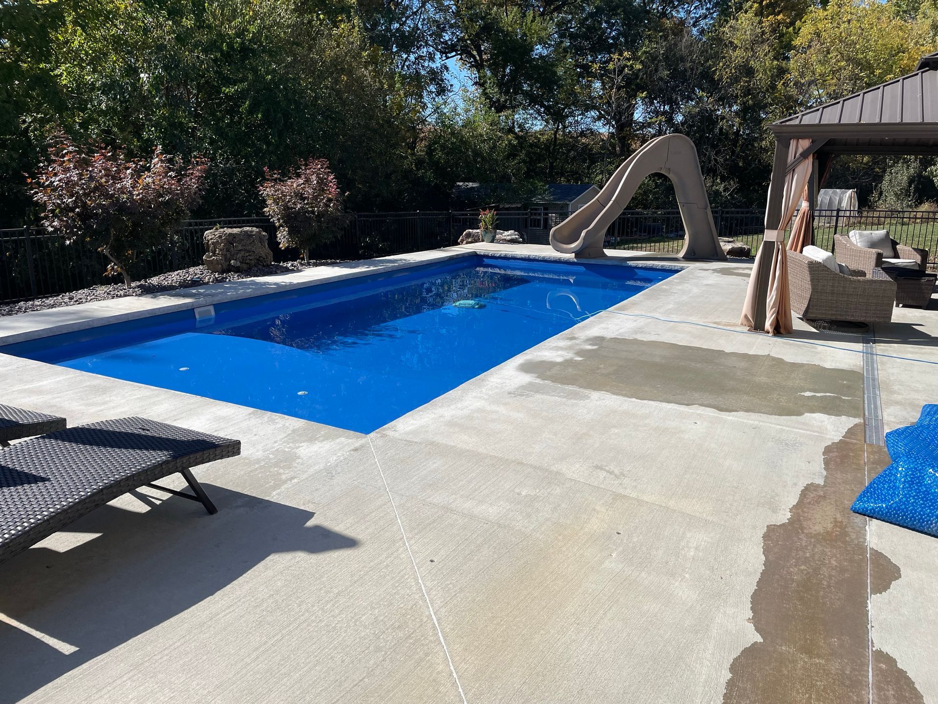 Rectangular pool with slide and patio seating. Blue water, concrete deck, brown pergola, trees, and sunlight.