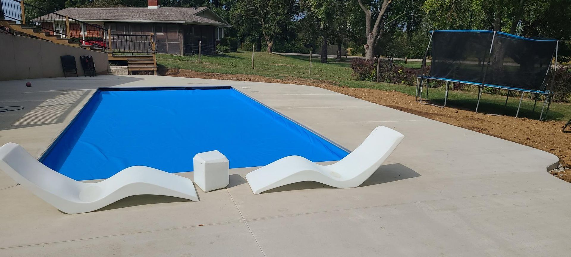 Swimming pool with blue cover, white lounge chairs, and a trampoline in the background.