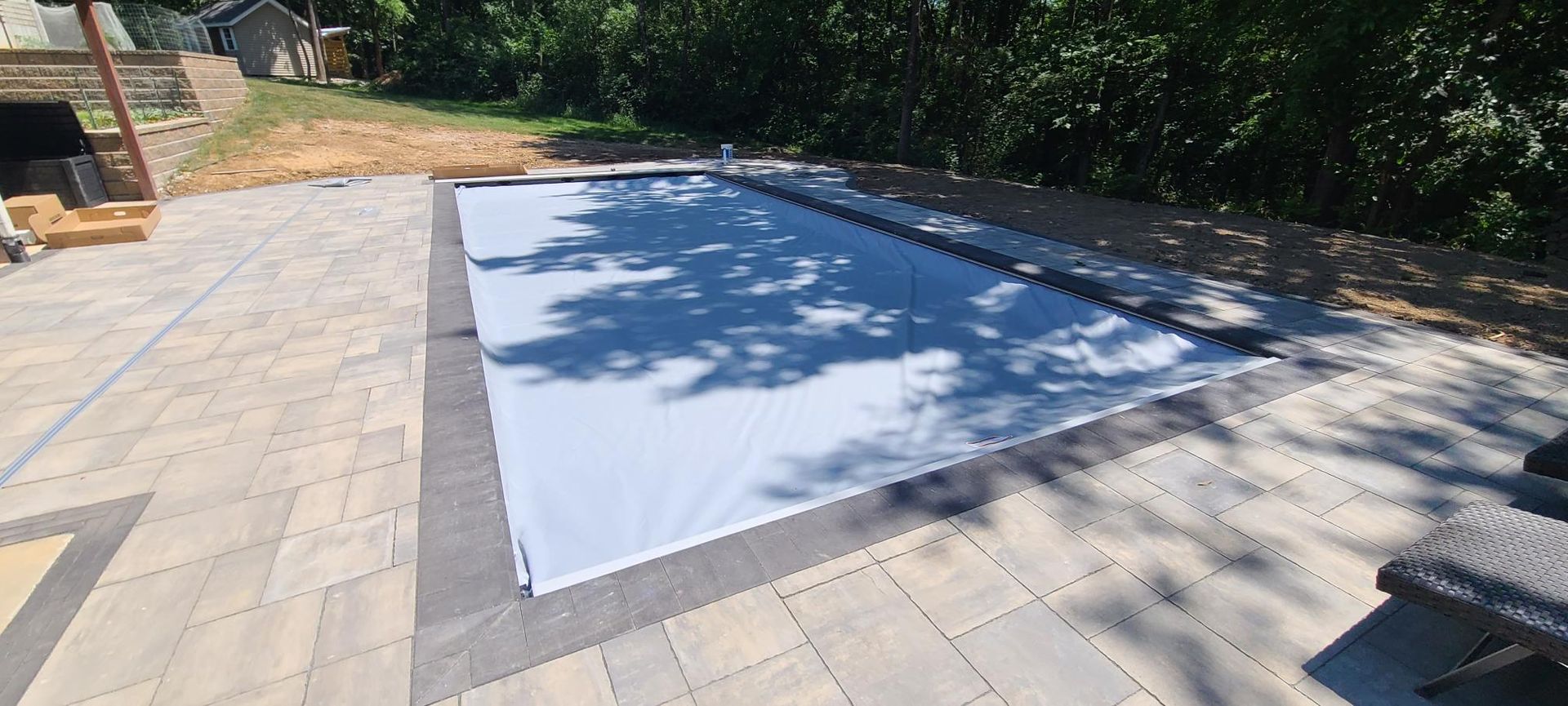 Pool covered by a light-colored tarp on a stone patio, surrounded by trees.