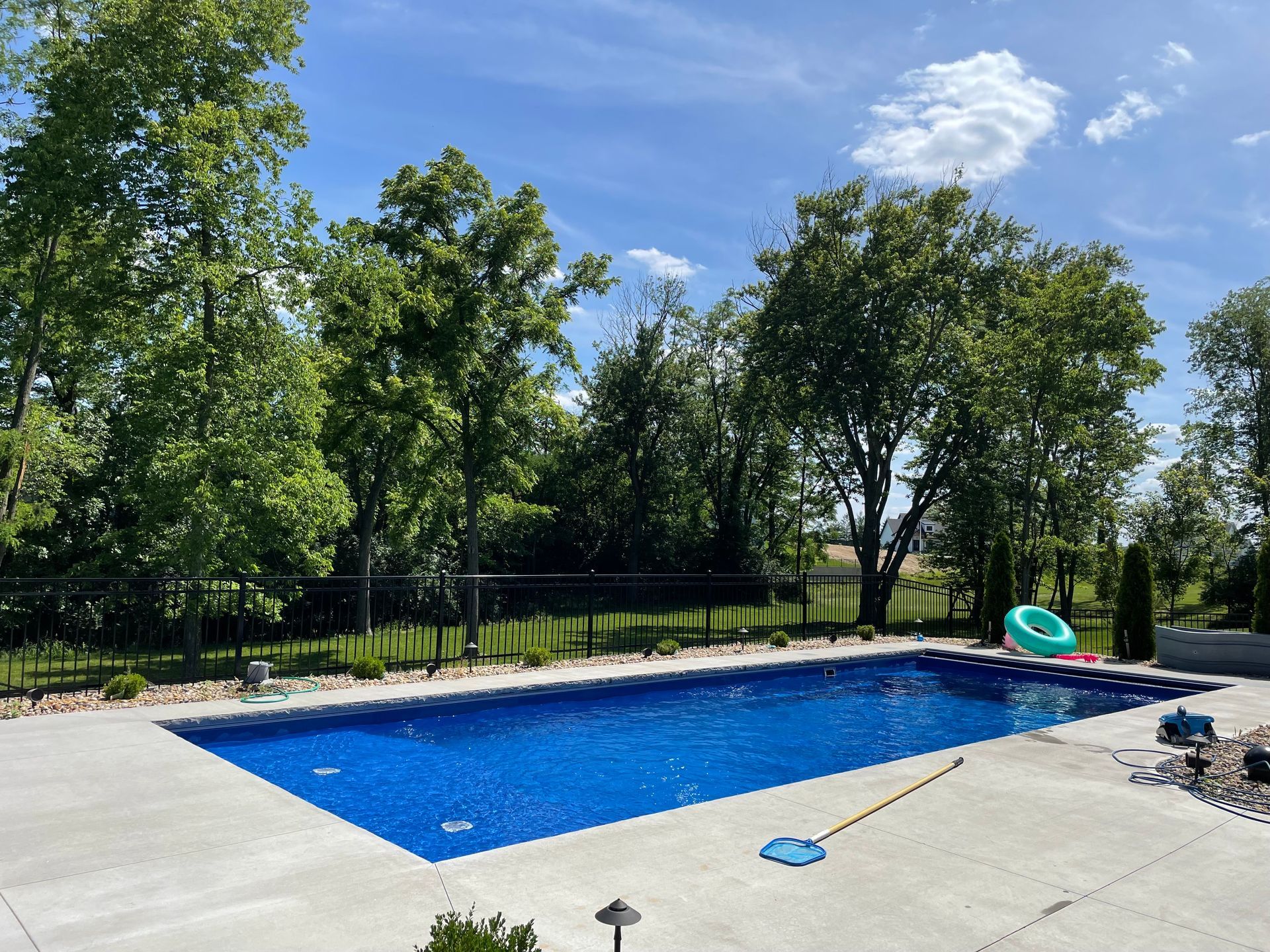 Blue rectangular pool surrounded by concrete, trees, and a blue sky with clouds.