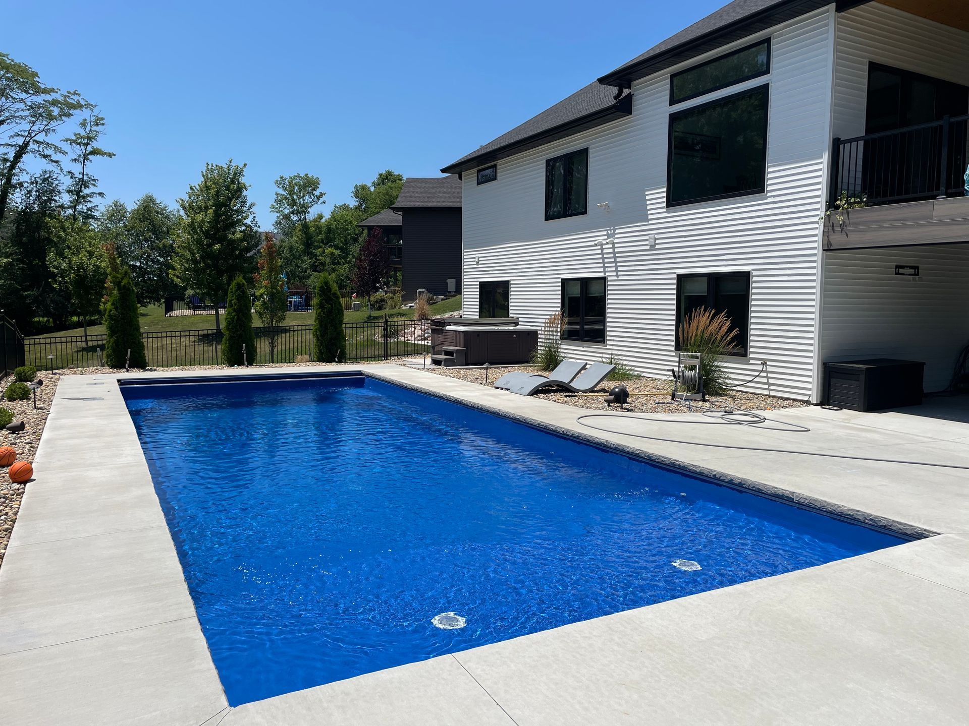 Rectangular blue pool surrounded by concrete patio, next to a modern white house on a sunny day.