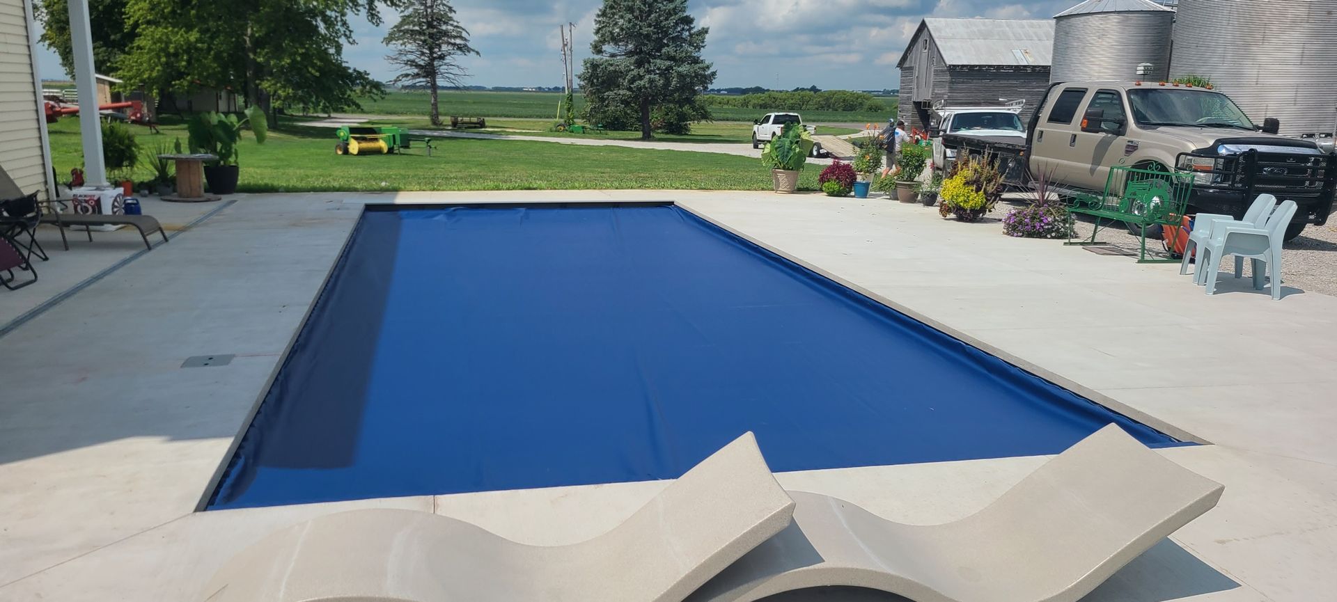 A rectangular blue pool covered with a tarp in a backyard. Concrete patio, plants, and vehicles in the background.
