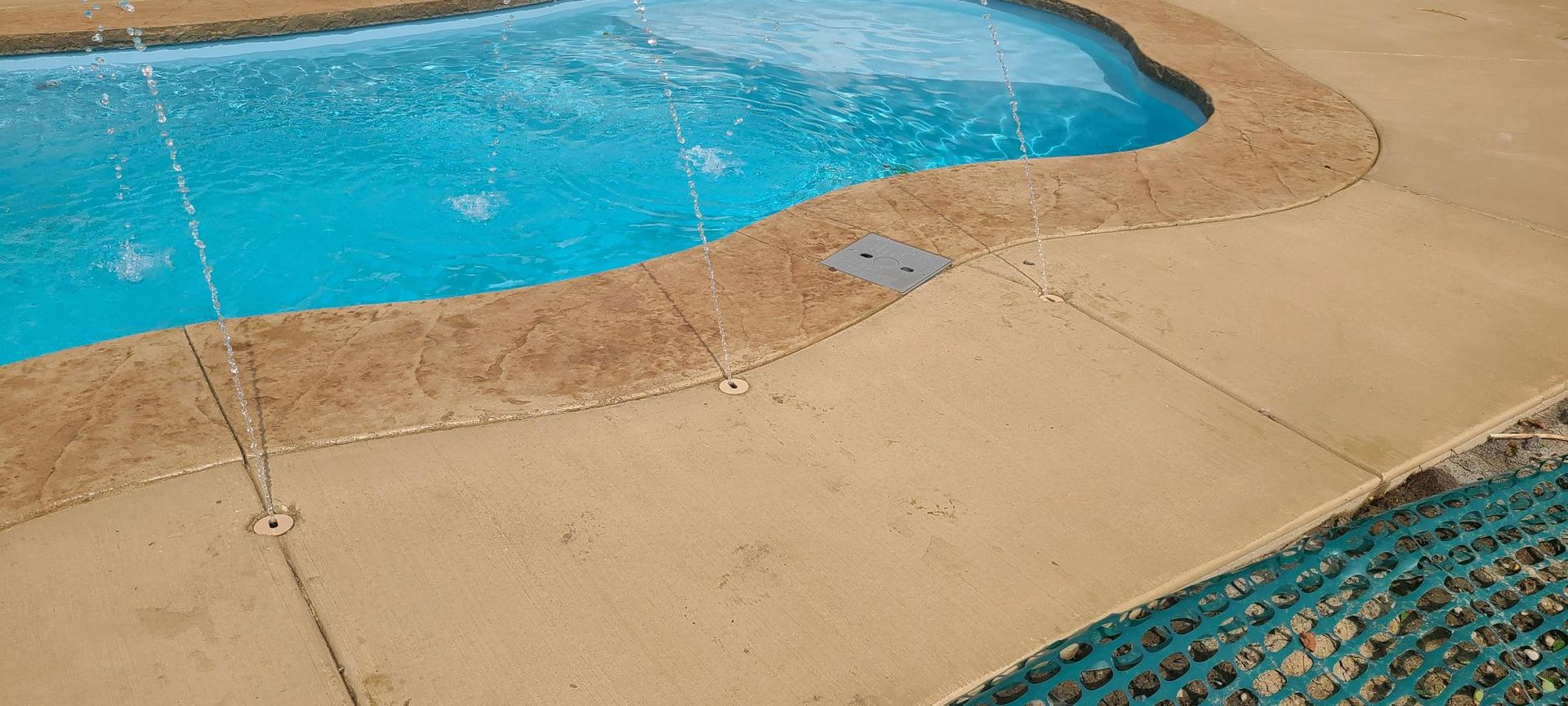 A turquoise pool with a brown stone border and a beige concrete deck.