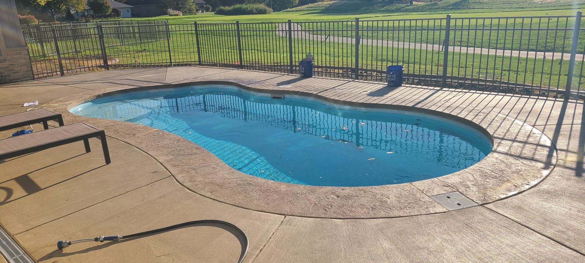 A small, kidney-shaped pool with a surrounding concrete patio and black metal fence. Blue water reflects sunlight.