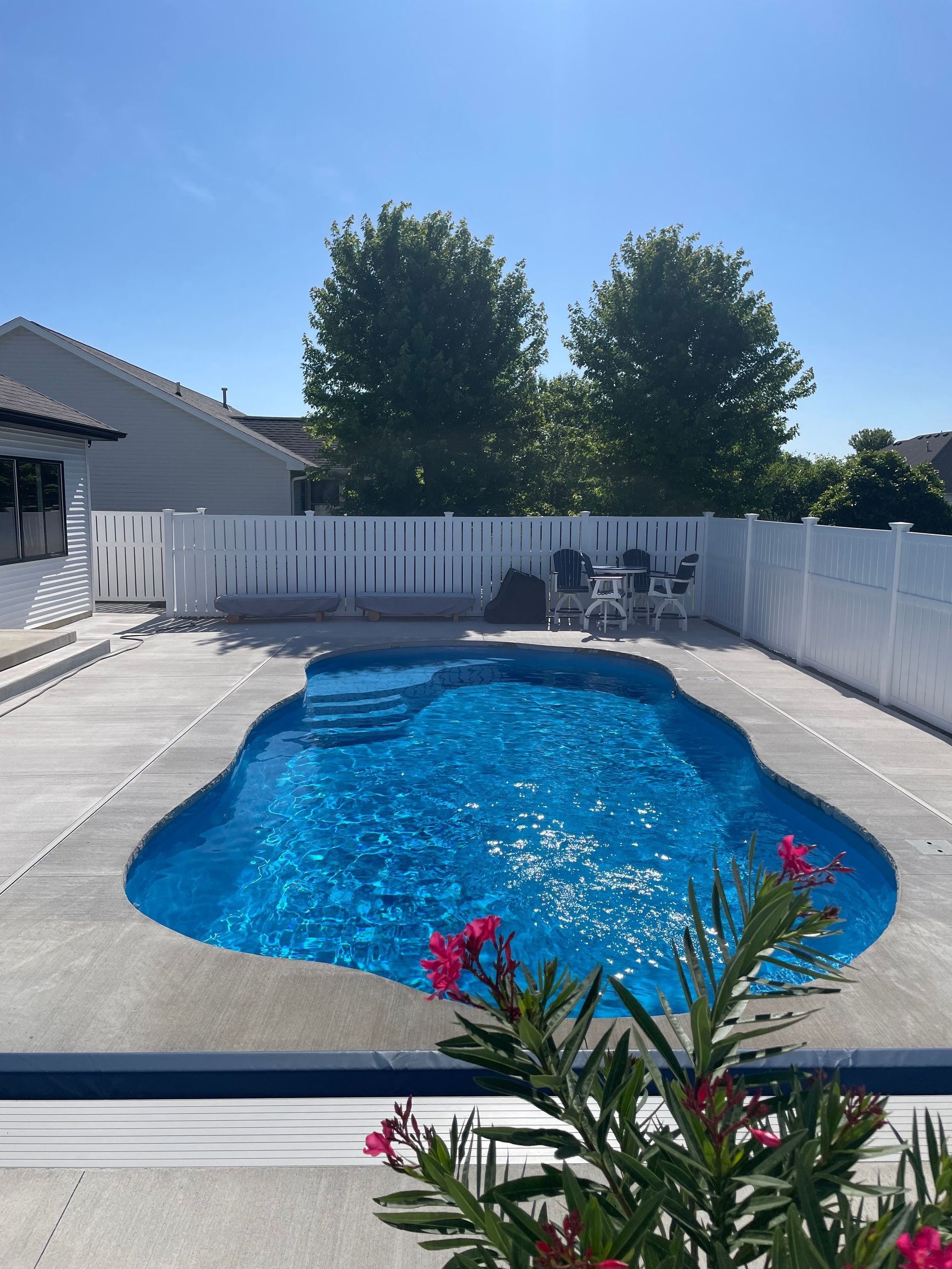 Blue pool in backyard, surrounded by concrete and white fence, under sunny blue sky.