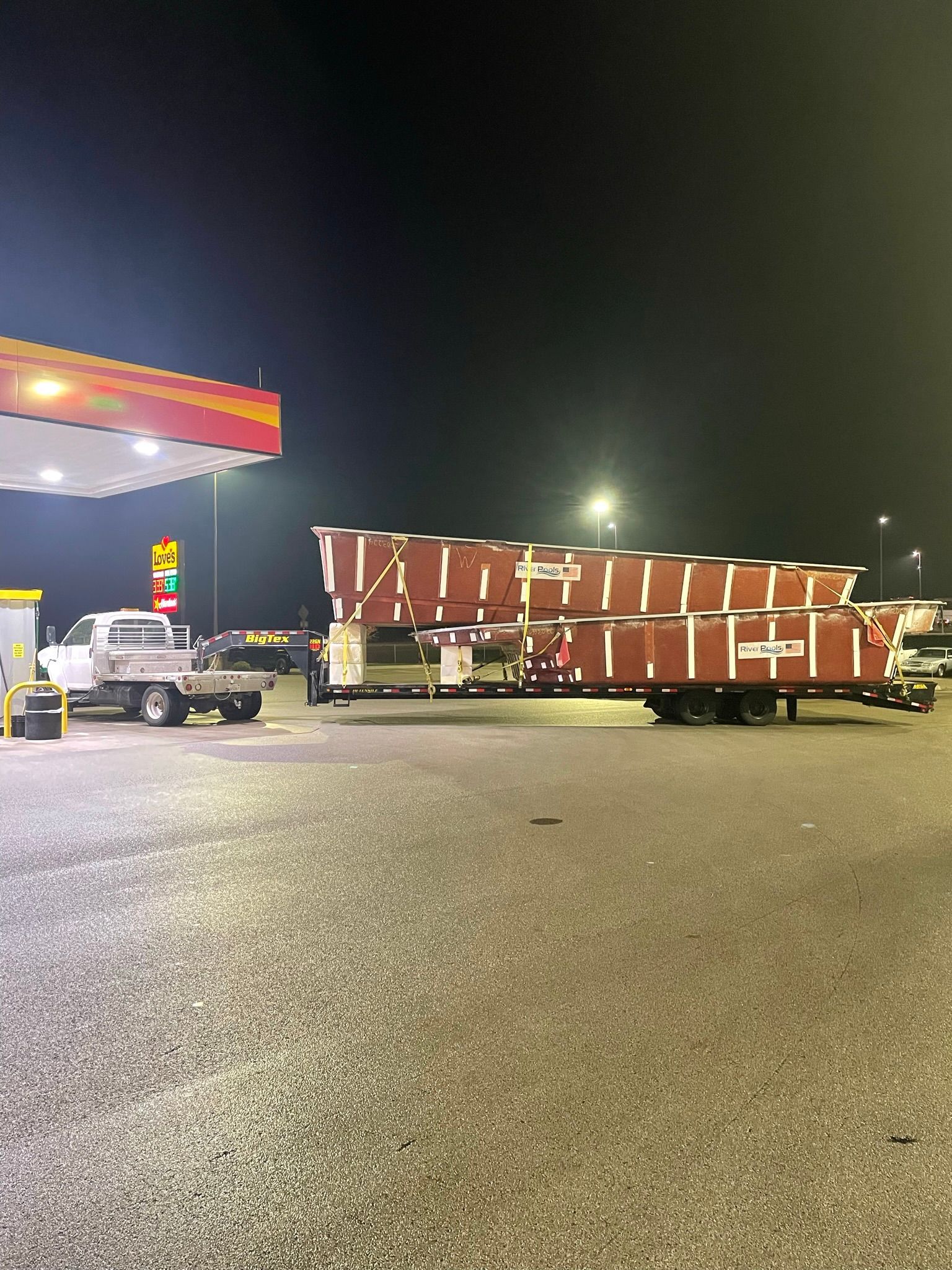 A truck with a trailer carrying several stacked, rectangular brown structures is parked at a gas station at night.