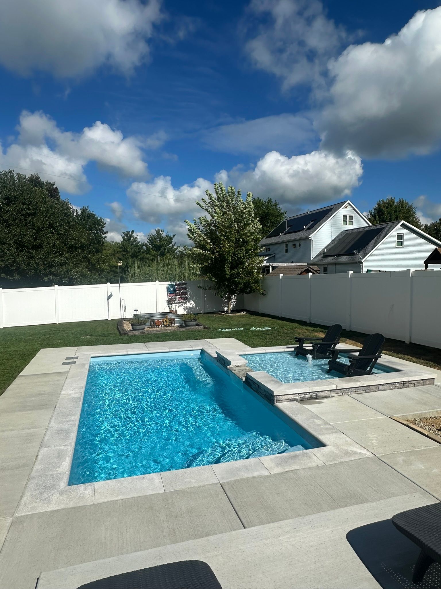 Backyard pool with spa, blue water, gray stone surround, white fence, blue sky, and a house in the background.