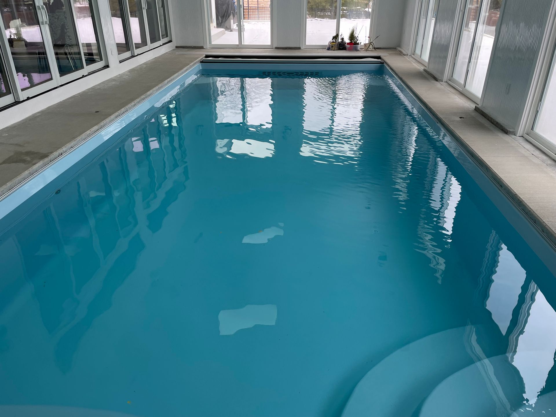 Indoor rectangular swimming pool with clear blue water and steps. Large windows on both sides.