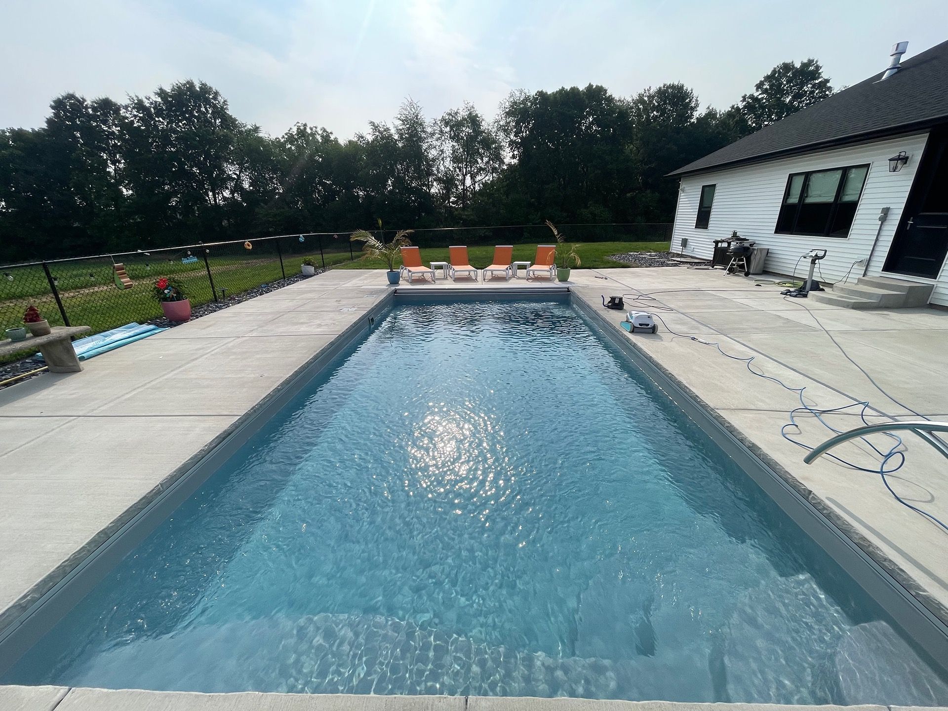 Rectangular pool with clear blue water, surrounded by concrete patio and lounge chairs. Sunny outdoor setting.