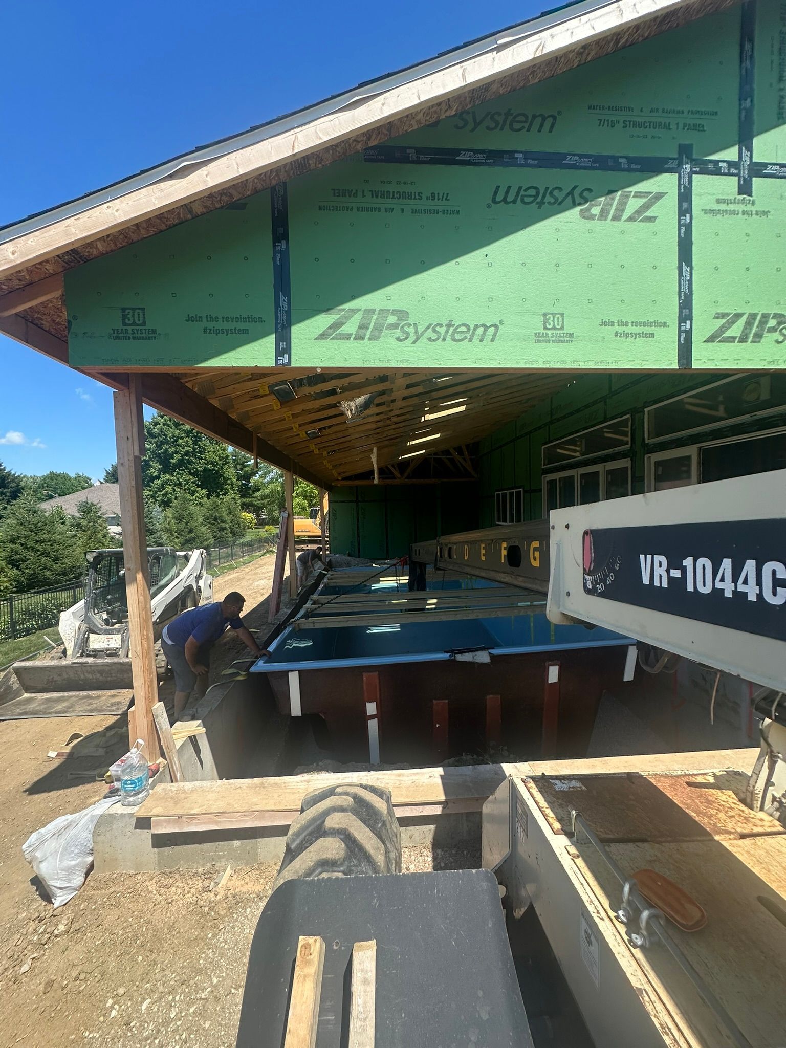 Construction site: workers near a covered structure with green sheathing. A small tractor is in the foreground.