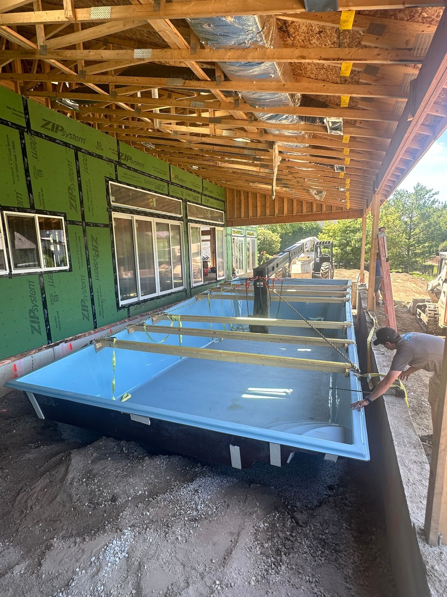 Pool construction under a covered structure, surrounded by dirt and unfinished building materials.