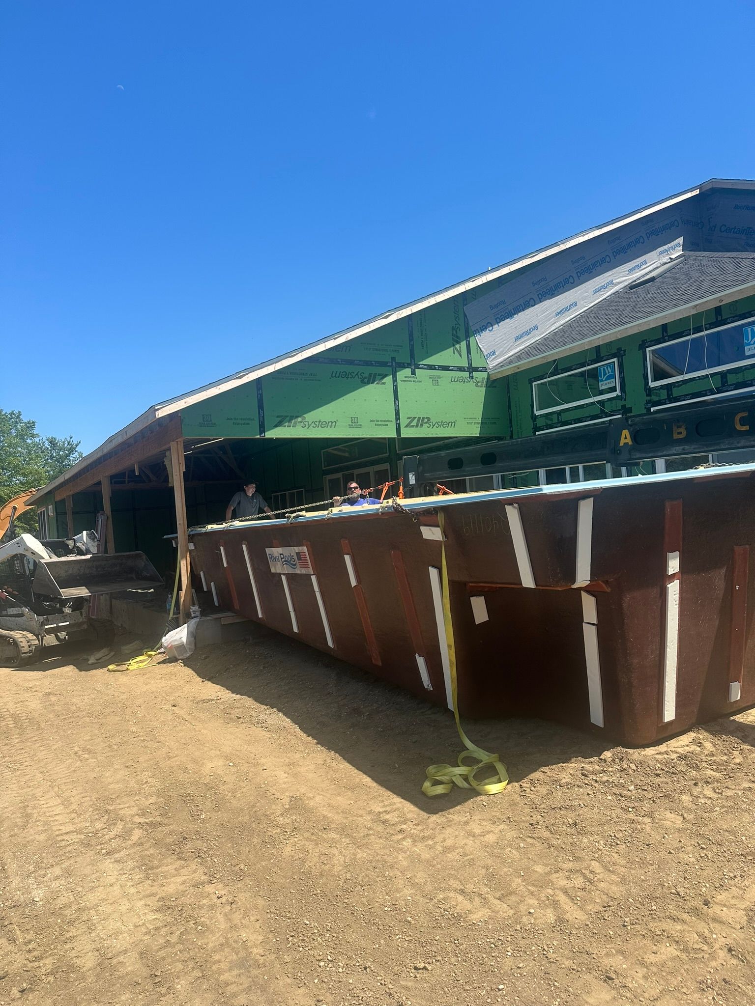 Construction site with a large brown dumpster in front of a building with green siding and a black roof