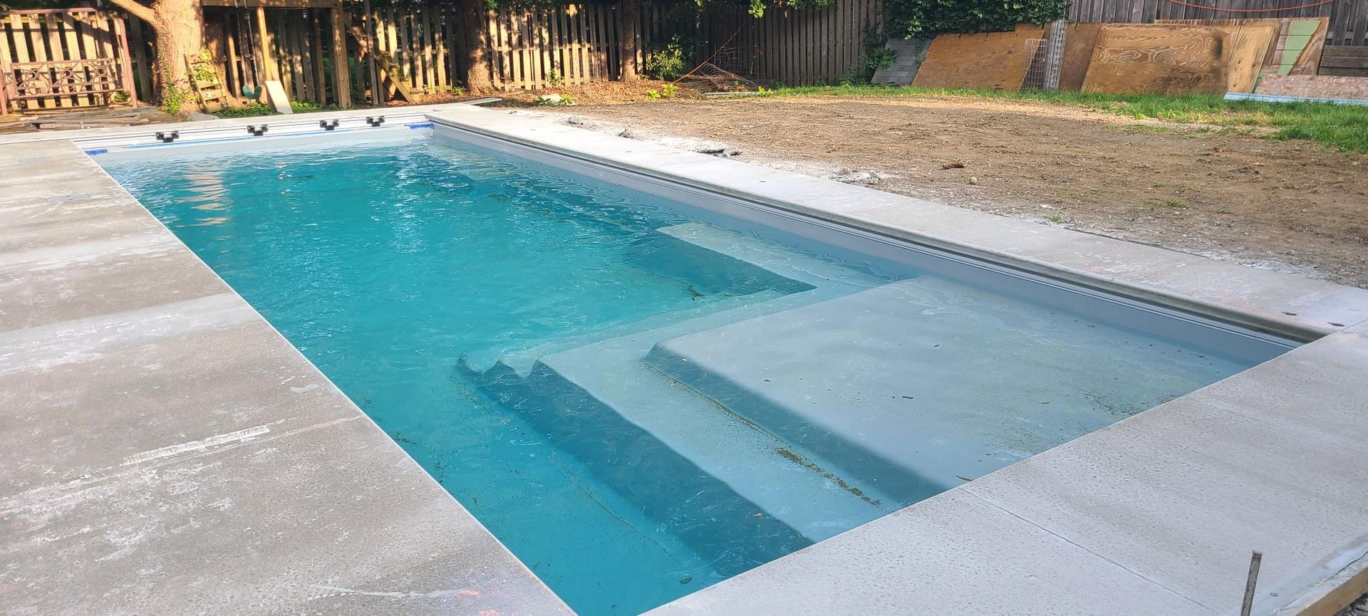 Rectangular swimming pool with steps, surrounded by concrete and grass. The water is clear blue.