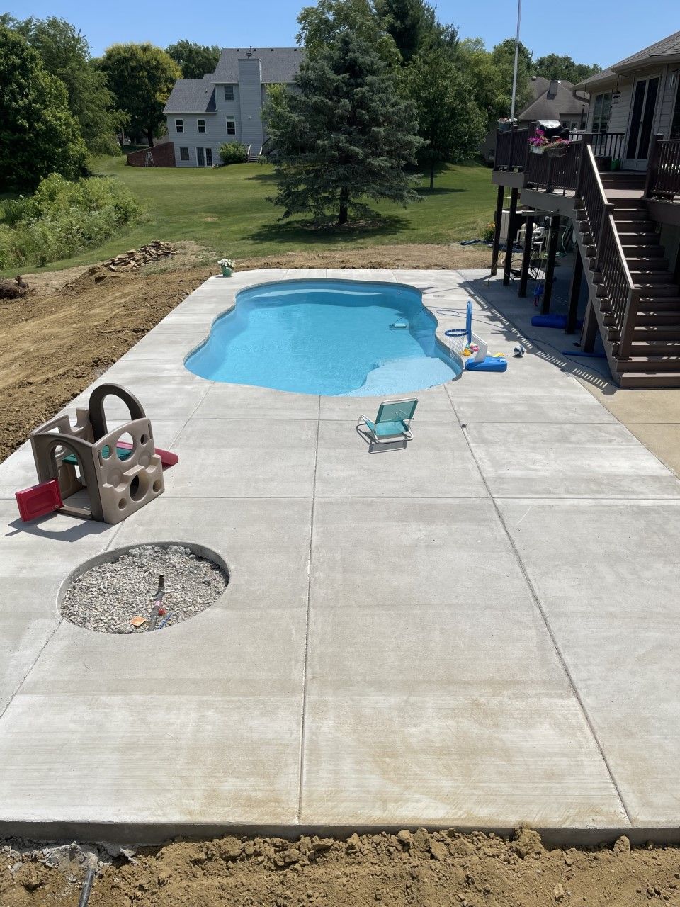 Concrete patio with a pool, fire pit, and play structure in a backyard setting.