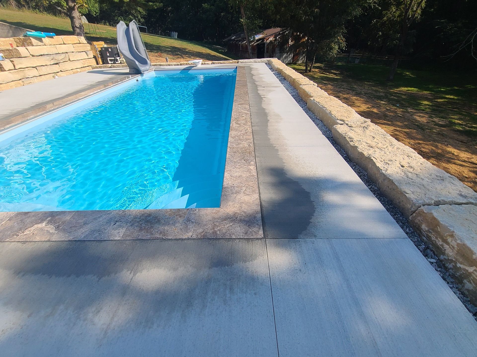 Swimming pool with blue water and gray concrete surrounding it, next to a stone wall.