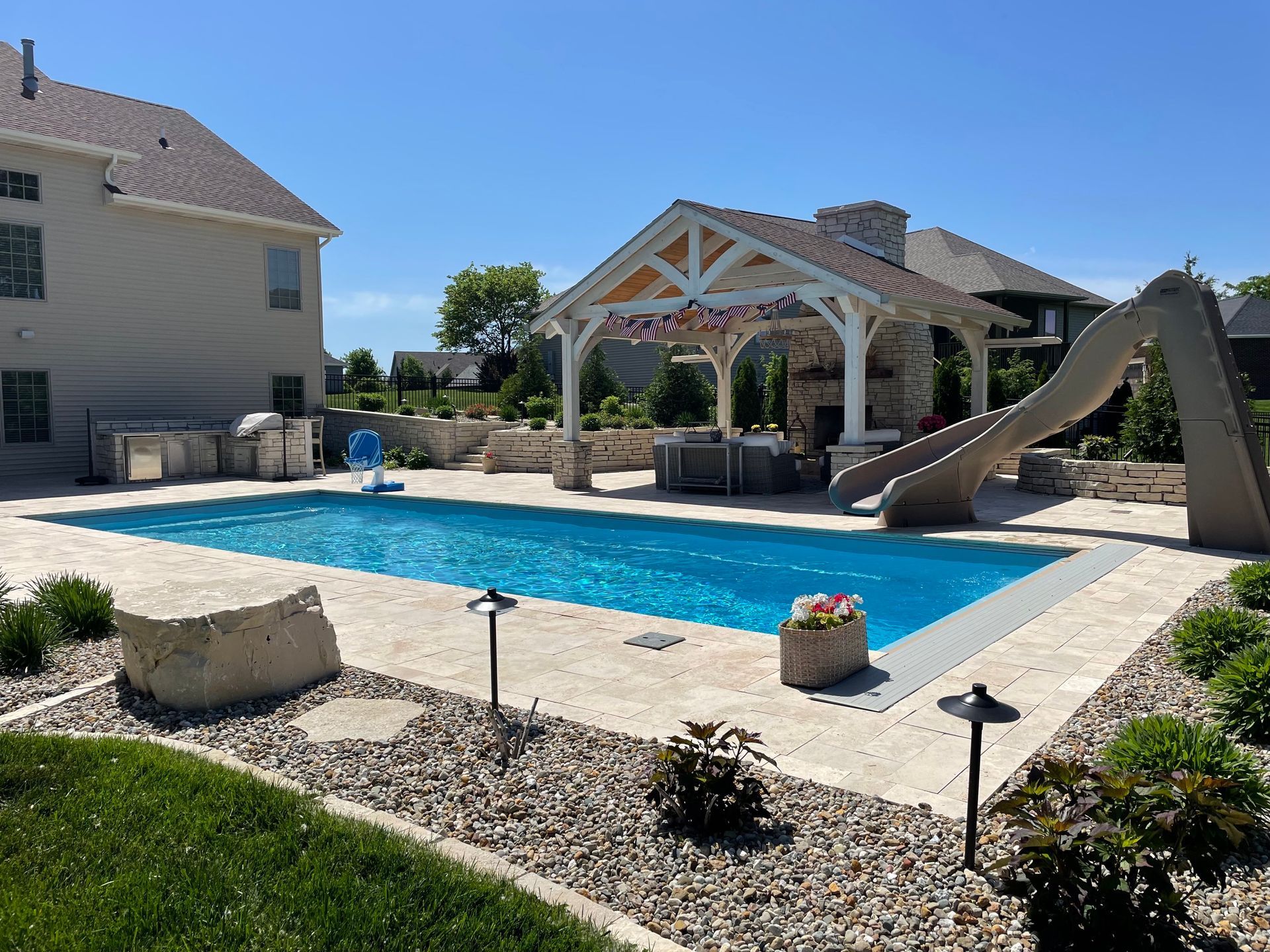 Swimming pool with slide, patio, and cabana in a backyard setting on a sunny day.