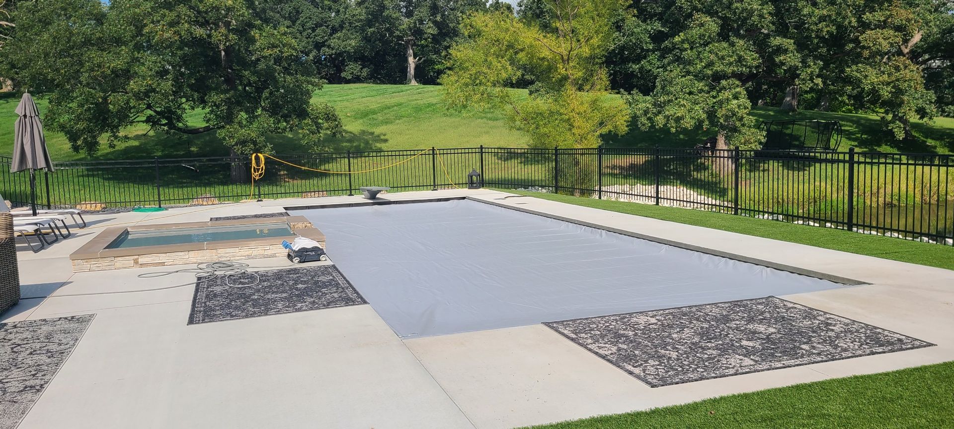 A backyard pool with a gray cover, surrounded by concrete and grass, with trees in the background.