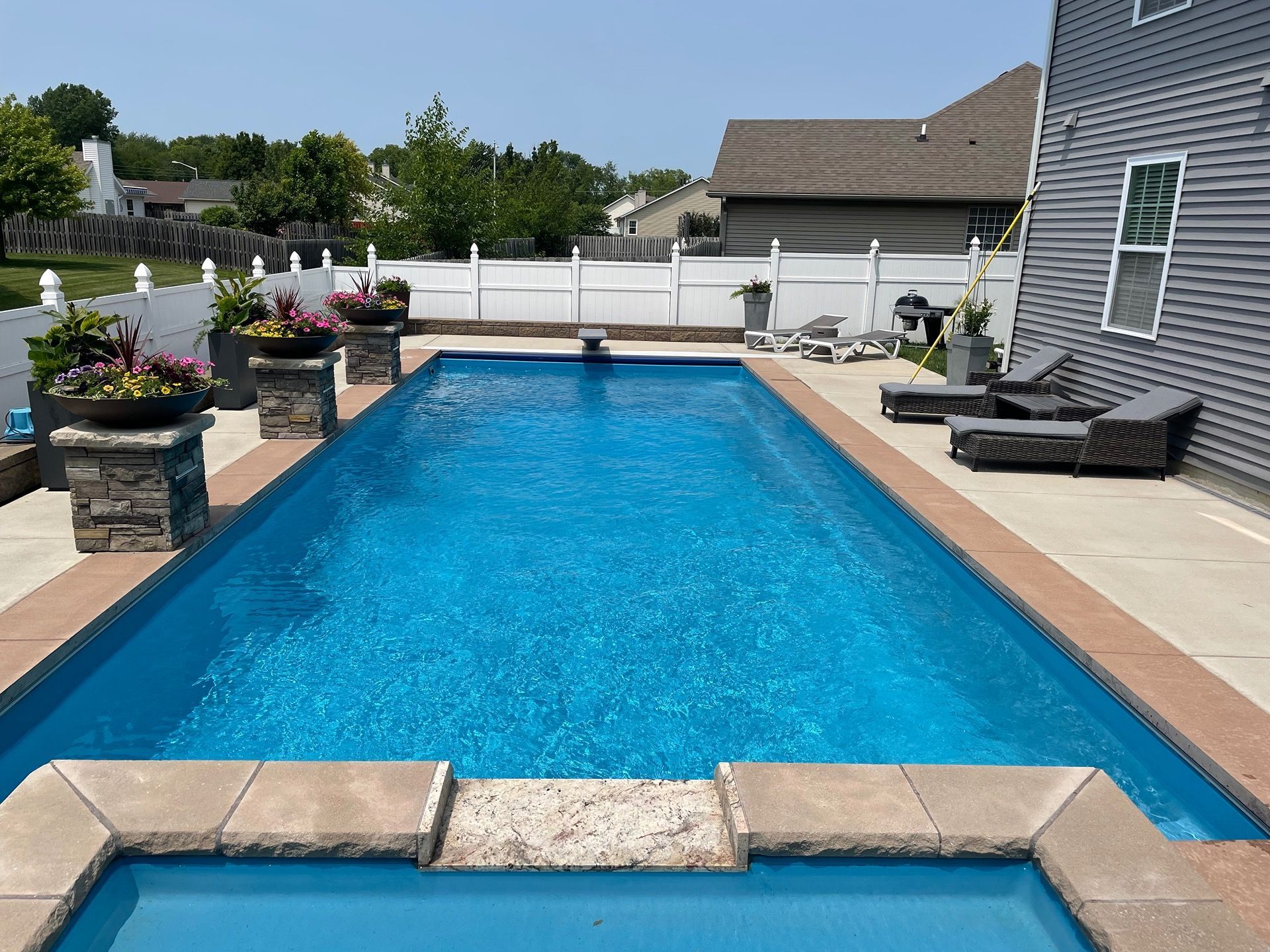 Rectangular blue swimming pool in a backyard, surrounded by a patio and a white fence.