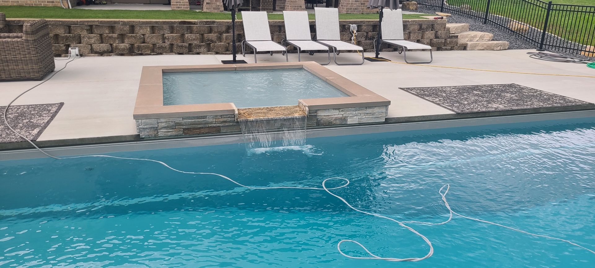 Pool with hot tub, lounge chairs, stone wall, and cascading water.